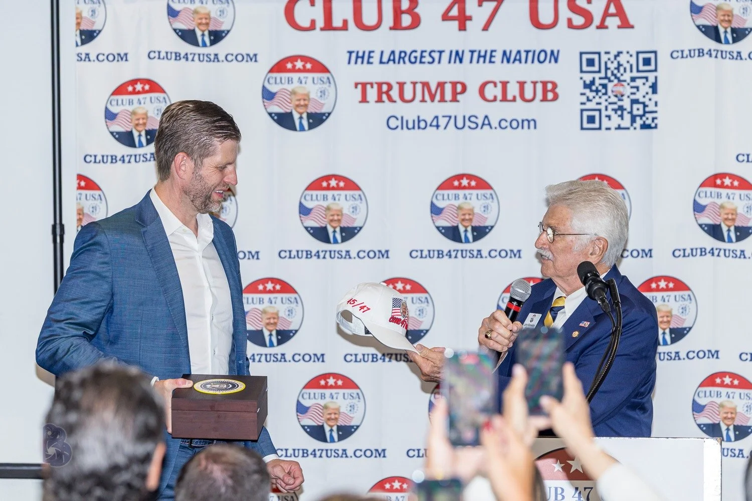 Two men at a political event, one older with white hair and glasses speaking into a microphone, the other younger in a blue suit, both smiling. The background displays a step and repeat banner with the logo for 'Club 47 USA' featuring an image of Don