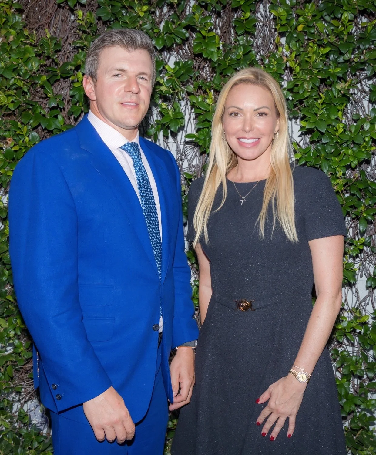 A man in a bright blue suit and a woman in a black dress standing in front of a leafy green wall, smiling at the camera.