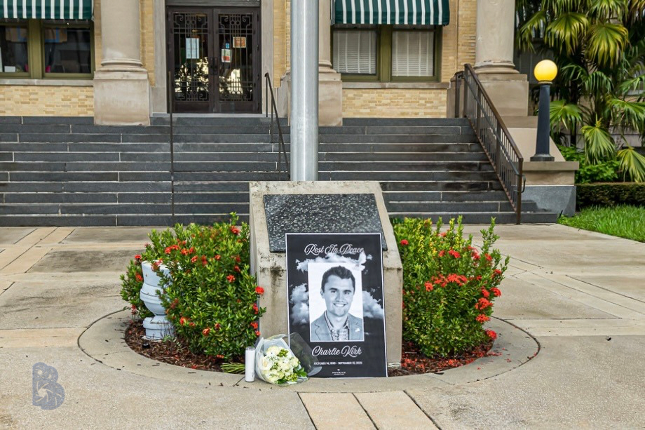 Memorial display with a black and white photo of a young man, surrounded by flowers, placed in front of a building with stairs, plants, and a lamppost.