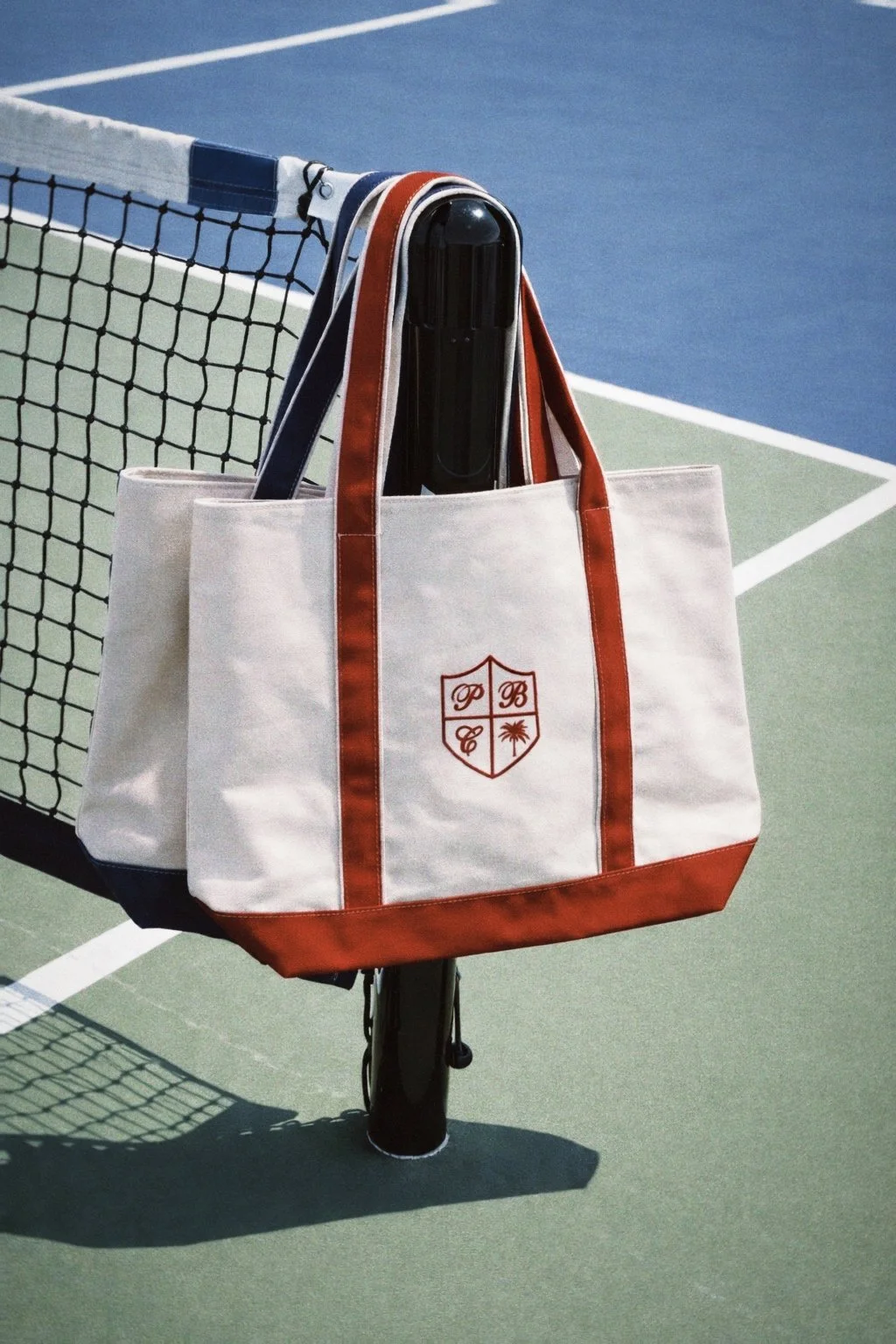 A white tote bag with red and blue accents hanging on a tennis net post on a tennis court. The tote has a crest with the initials P B C and a palm tree.