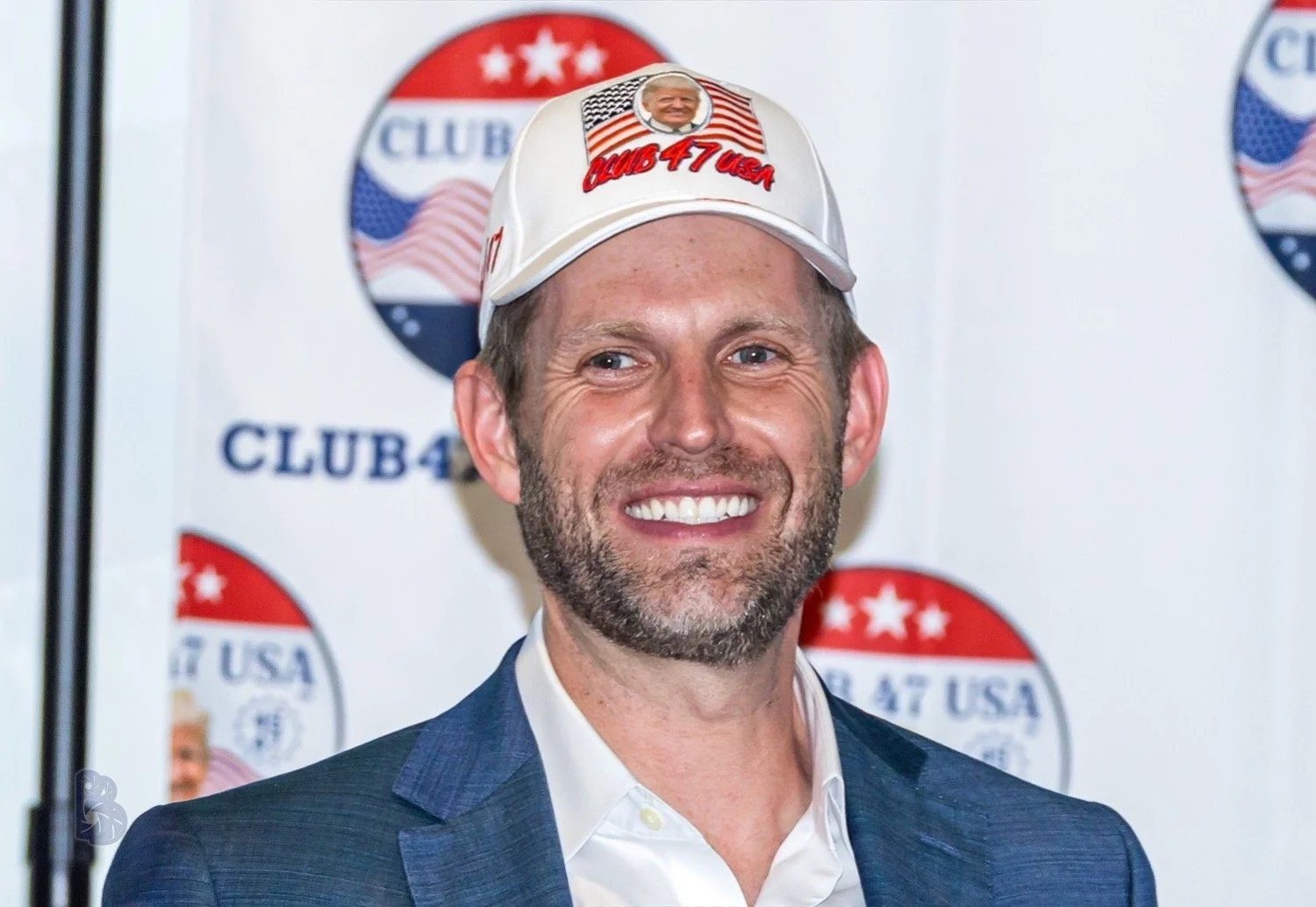 A smiling man with a beard and blue eyes, wearing a white baseball cap with patriotic and American flag graphics and the words 'Club 47 USA,' is standing in front of a white background with circular logos that include American flags and the words 'Cl