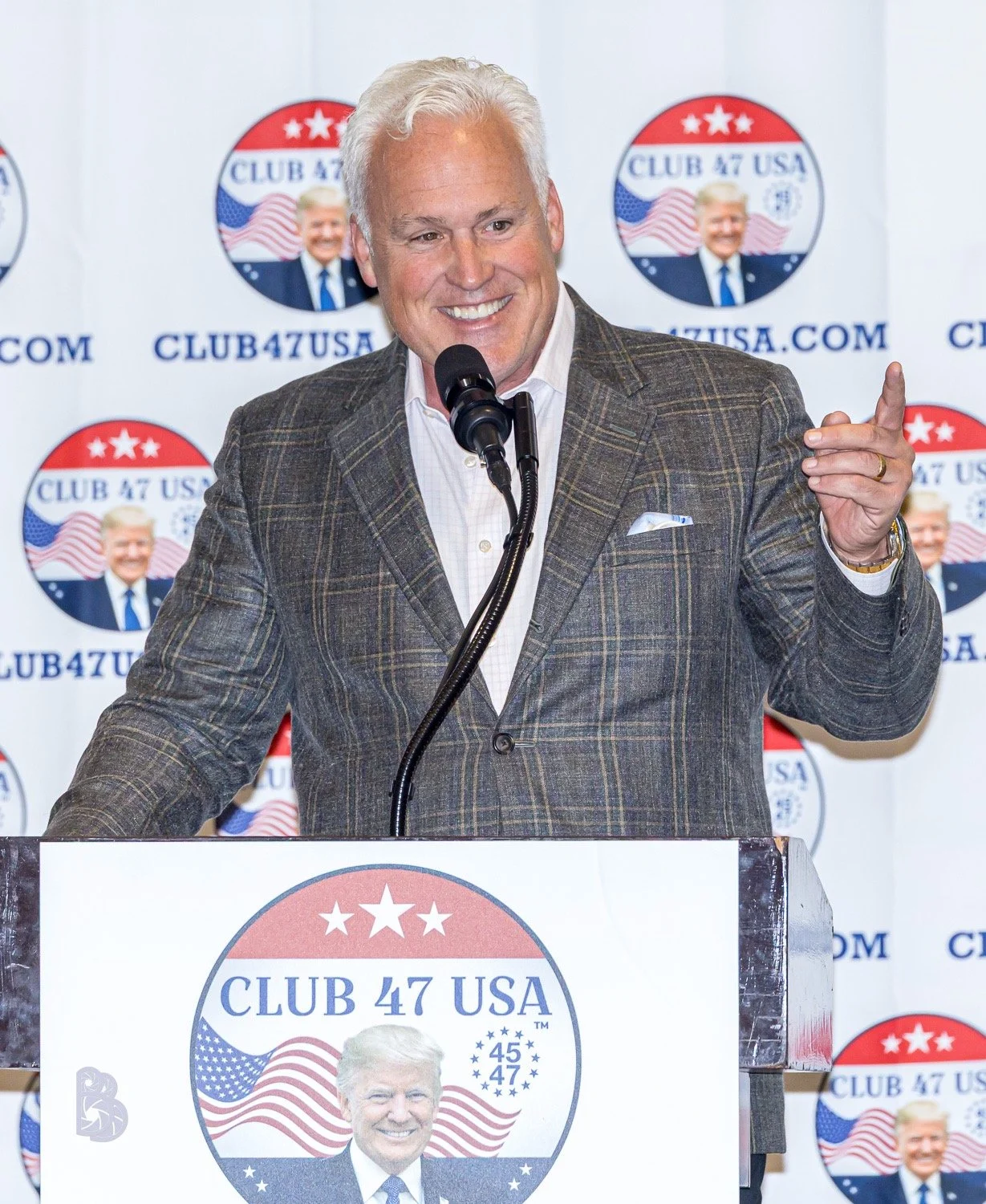 Man with white hair and a checkered jacket speaking at a podium with a microphone, smiling, in front of a backdrop with 'CLUB 47 USA' logos featuring former President Donald Trump.