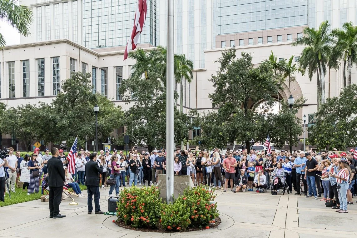 A crowd gathering outdoors near a flagpole in an urban park with tall buildings in the background, some people holding American flags, and a speaker addressing the crowd.