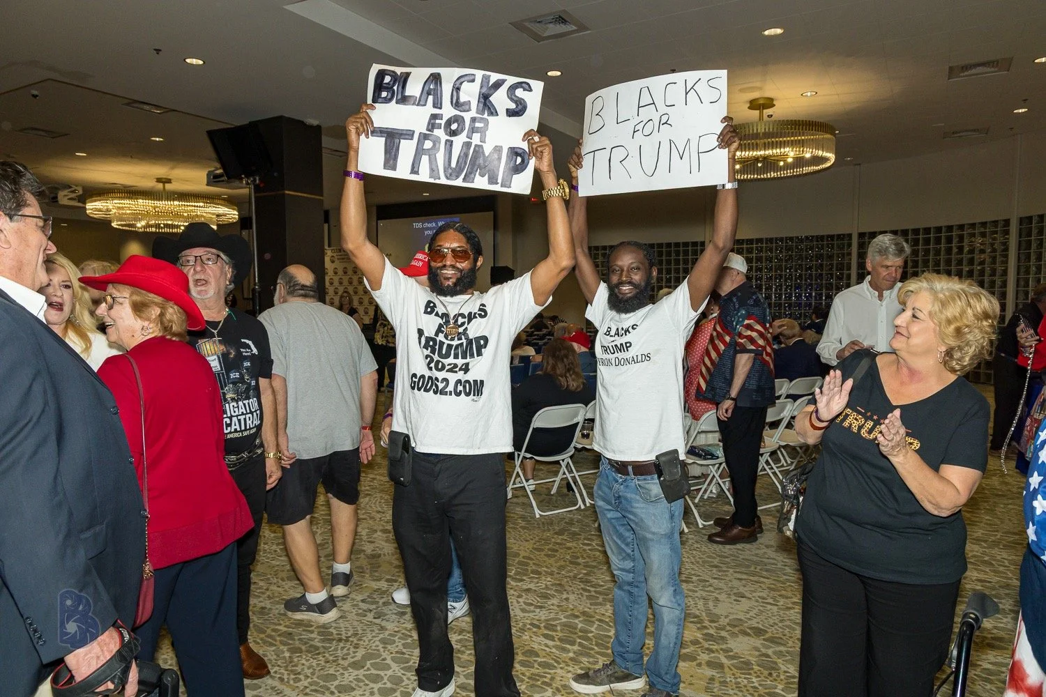 Two men at a gathering holding signs that say 'Black for Trump' with one sign also mentioning '2024'. Other people are around them, some smiling and clapping, in an indoor setting with chairs and chandeliers.