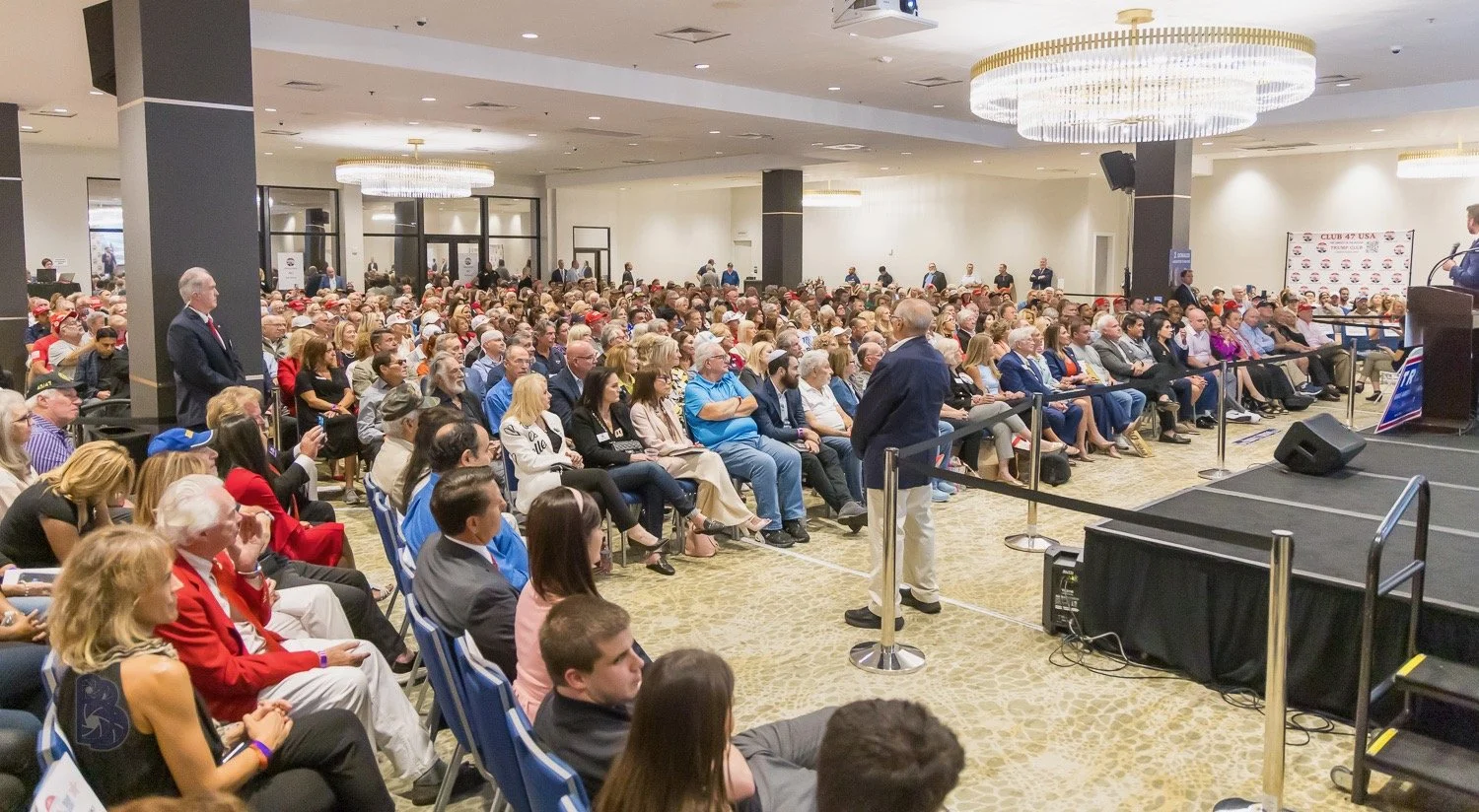 A large indoor conference with many attendees seated facing a stage, where a speaker is addressing the audience.
