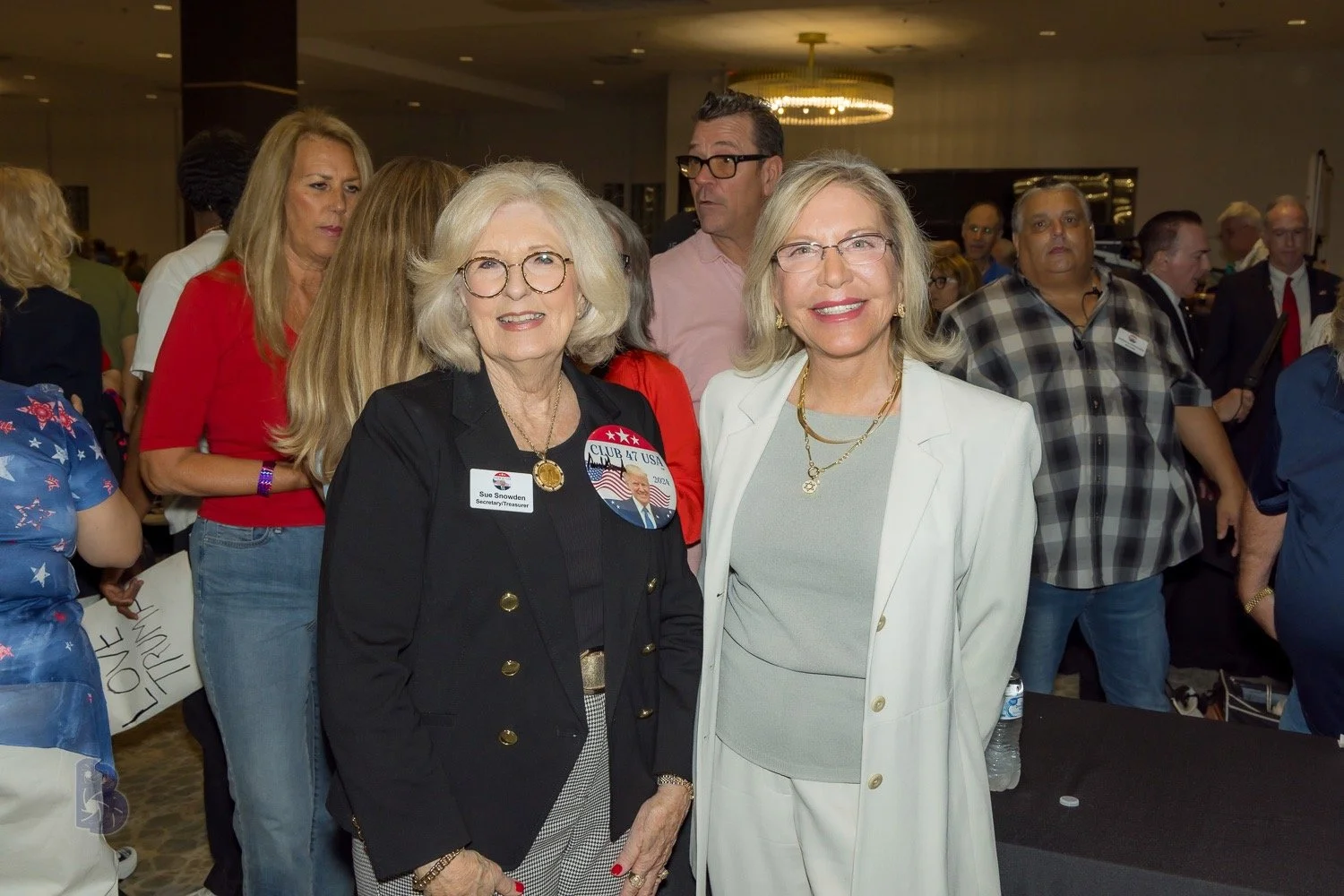Two women smiling at a political event, surrounded by a crowd of people.