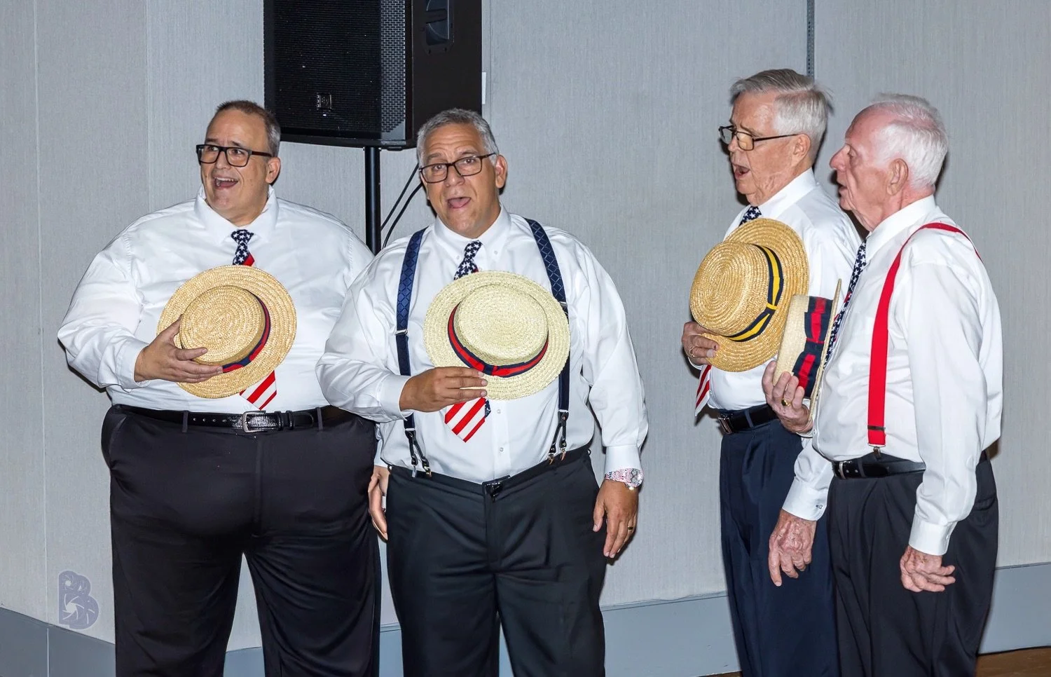 Five elderly men dressed in white shirts, black pants, and patriotic ties, holding straw hats with red, white, and blue ribbons, are standing indoors and singing.