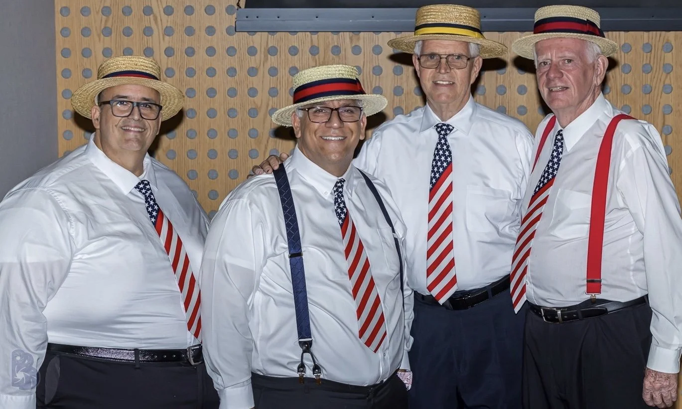 Five older men dressed in white shirts, patriotic ties, suspenders, and straw hats with red, white, and blue bands, standing together and smiling at an indoor event.