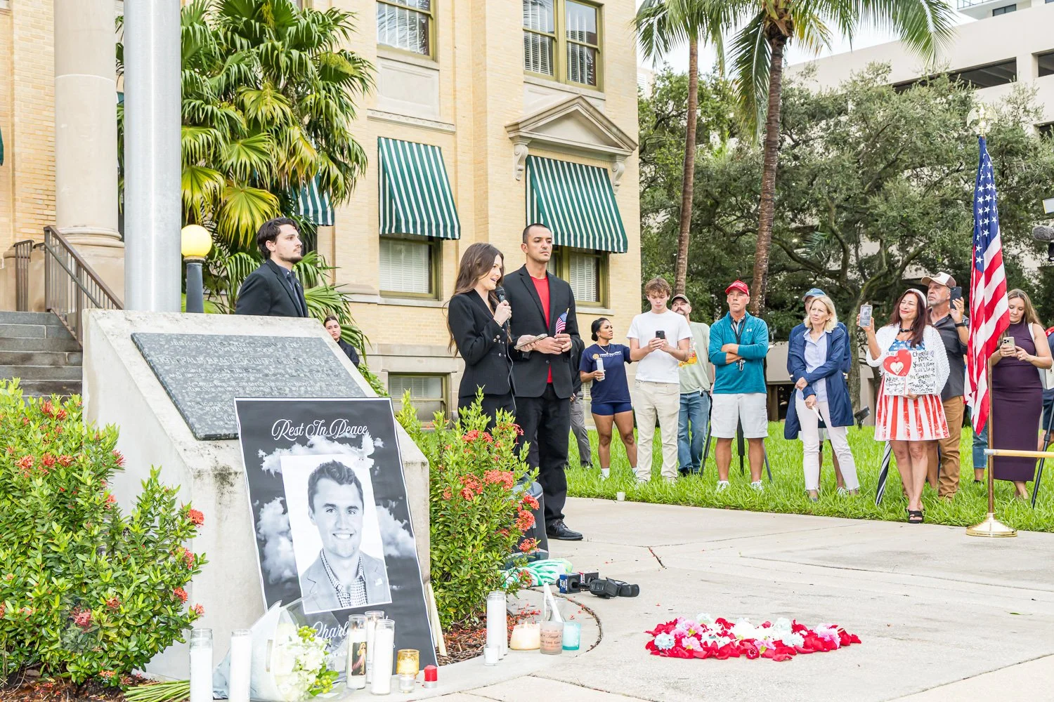 People gathering at a memorial site outdoors, paying respects with flowers and candles for a young man whose large black-and-white photo is displayed. A woman is speaking into a microphone, and others are holding phones or signs. A large American fla