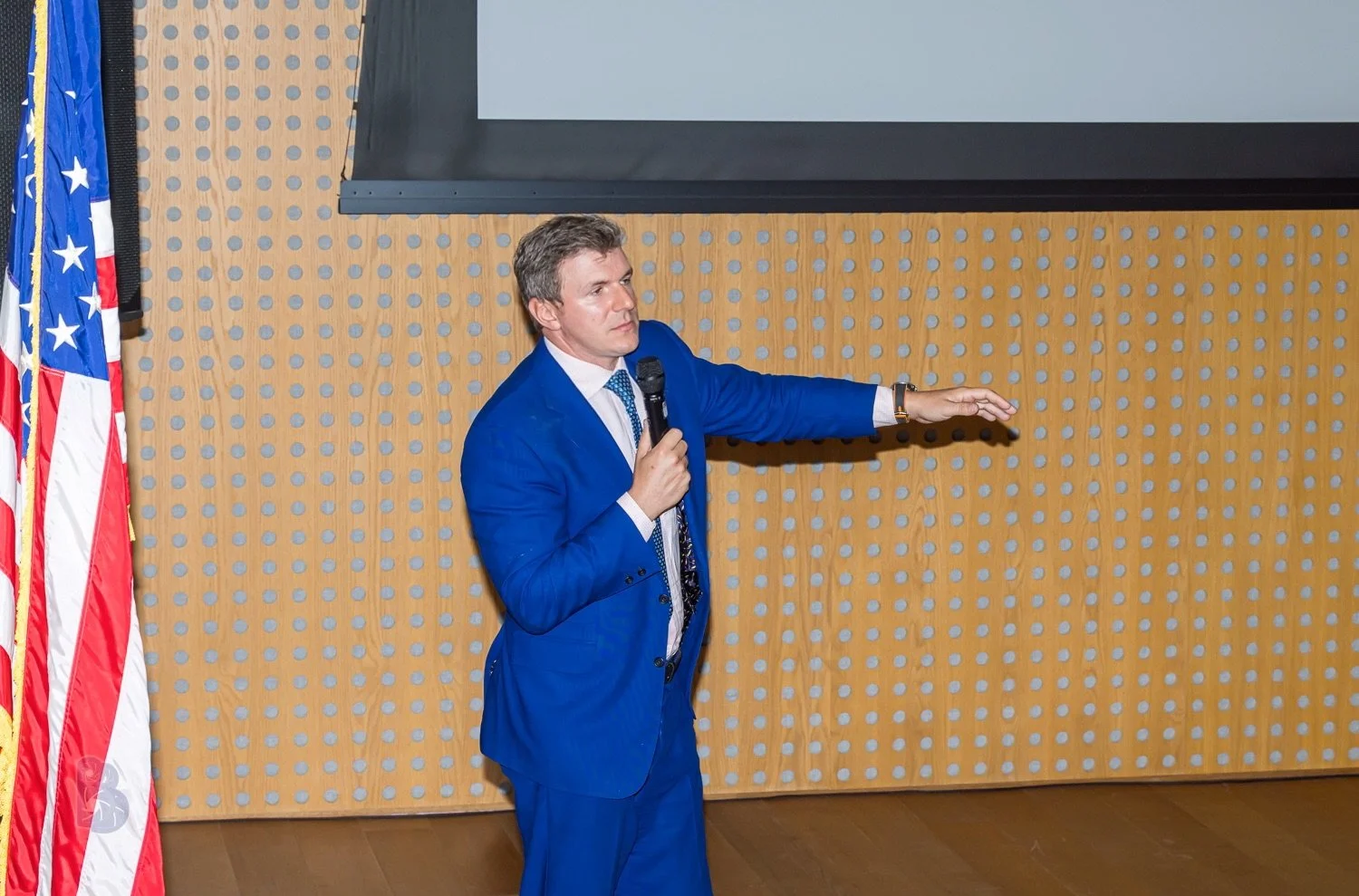 A man in a blue suit holding a microphone and gesturing to a presentation screen, standing beside American flags.