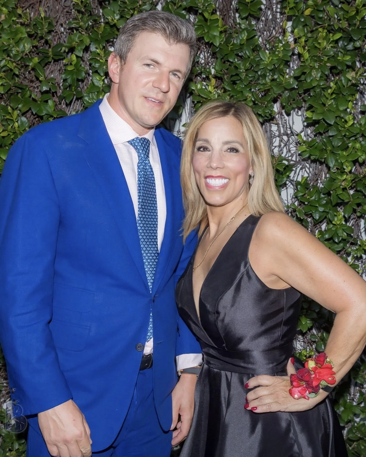 A man in a blue suit and a woman in a black dress standing outdoors against a leafy green background, smiling at the camera.