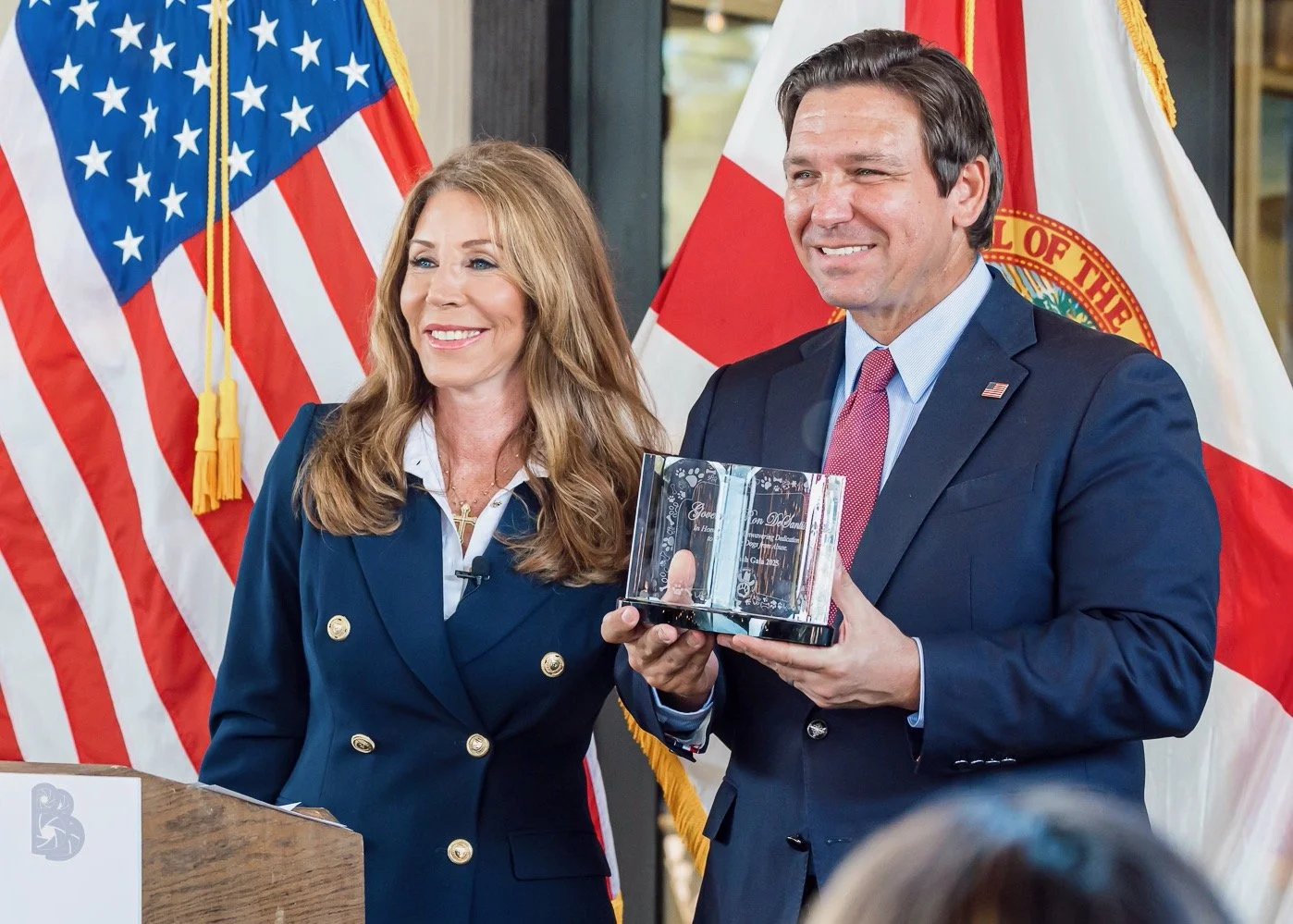 A man and a woman smiling, standing in front of American and Florida flags. The man is holding a clear glass plaque or award, and the woman is wearing a dark blue blazer with gold buttons.