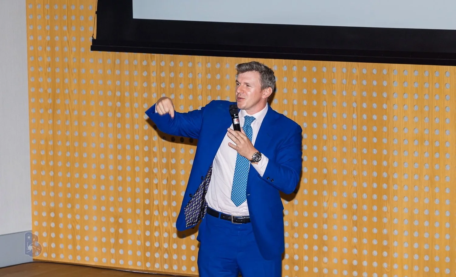 A man in a blue suit and blue tie giving a presentation or speech in front of a yellow wall with a black screen above.