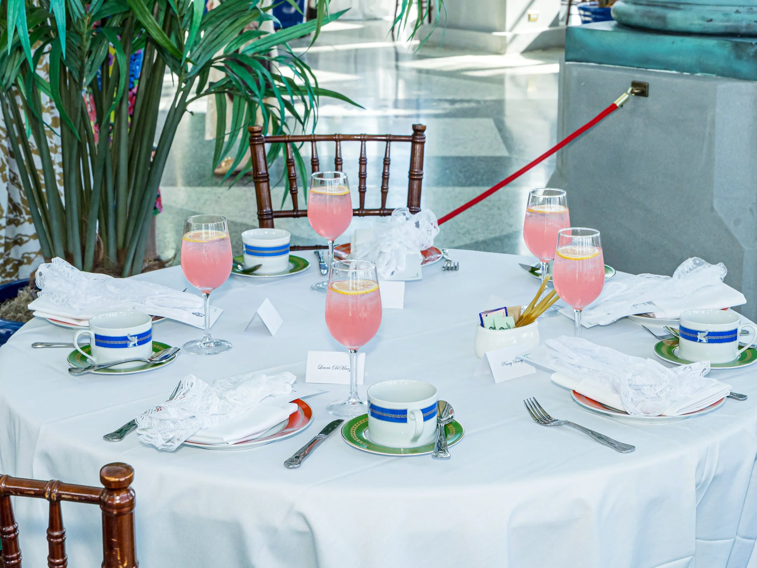 A round table set for a meal with six place settings, each with a cup, saucer, plate, and utensils. There are six pink cocktails with lemon slices on top, glasses, and a white napkin at each place. The table has a white tablecloth and is decorated wi