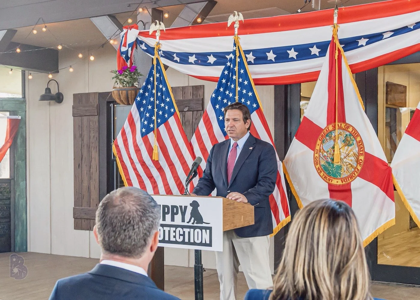 A man in a suit giving a speech at a podium decorated with American flags and a Florida state flag, with people seated in front, during a patriotic event.