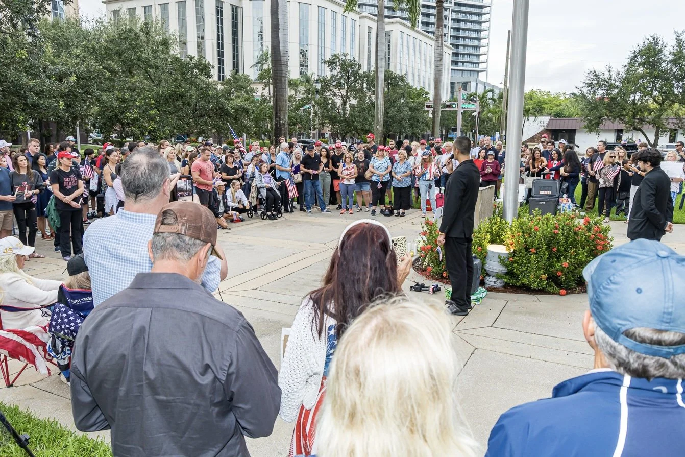 People gather outdoors for a rally or vigil near a flagpole with American flags, with some individuals holding flags and signs, and two men dressed in black standing in front addressing the crowd.