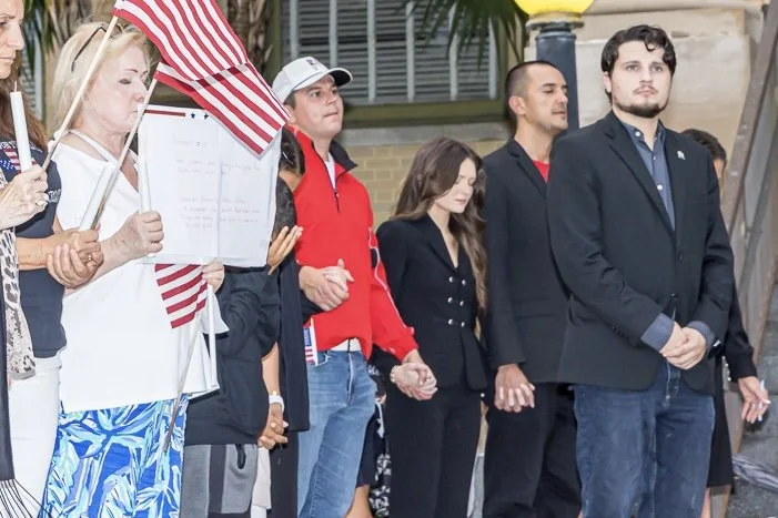 People standing in a line, holding hands, with some holding American flags, during a public gathering or memorial event.