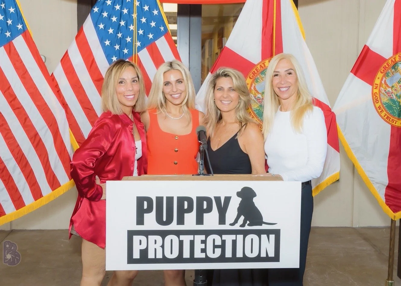 Four women standing behind a podium with a sign that says "Puppy Protection," in front of American and Florida flags, smiling at the camera.