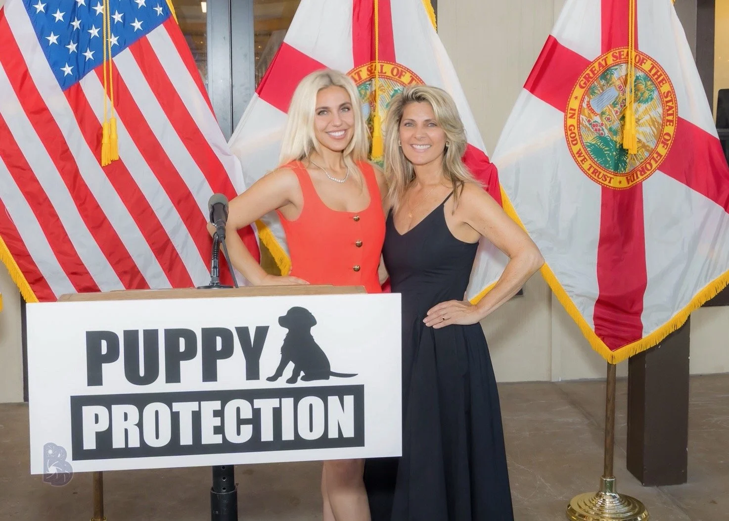 Two women standing in front of American and Florida flags at a puppy protection event, with a sign in front of them that reads 'PUPPY PROTECTION' and features a silhouette of a puppy.