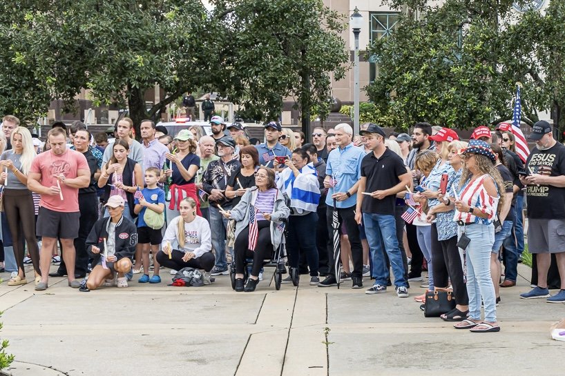 A crowd of Americans gathered outdoors for a patriotic event, some holding small American flags and wearing patriotic attire and hats.