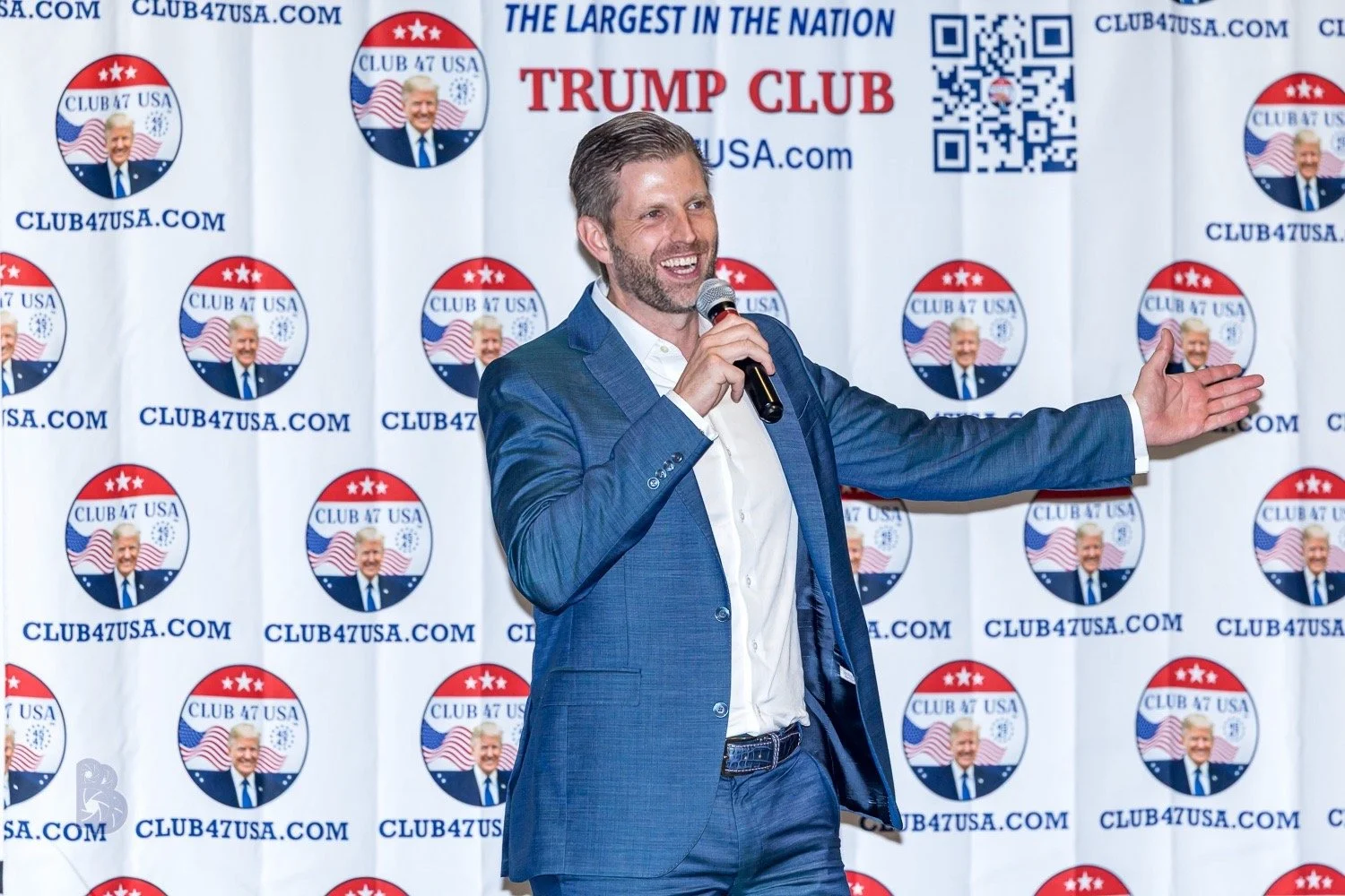 A man in a blue suit and white shirt holding a microphone and gesturing with his left hand, standing in front of a backdrop with logos for 'Club 47 USA' and 'Trump Club'. The backdrop has repetitive circular logos with an older man's face, American f