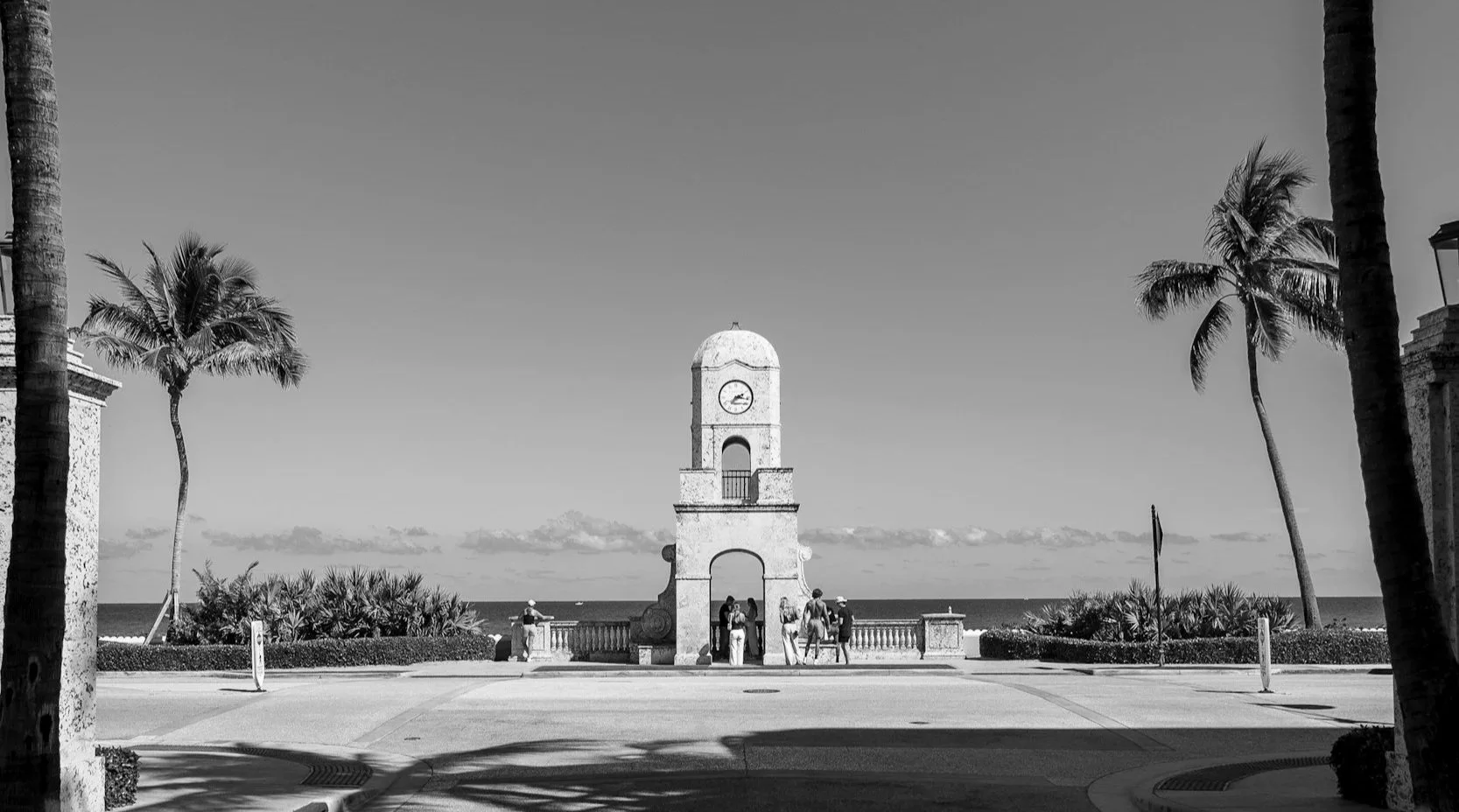 A seaside promenade with a clock tower in the center, surrounded by palm trees and a view of the ocean in the background, in black and white.