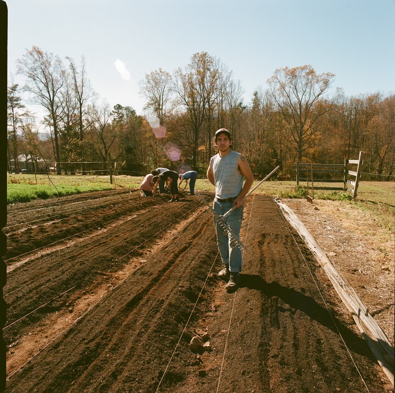 A man holding a rake standing on a freshly tilled garden bed with a group of people working in the background. The scene takes place outdoors on a sunny day with trees in the distance.