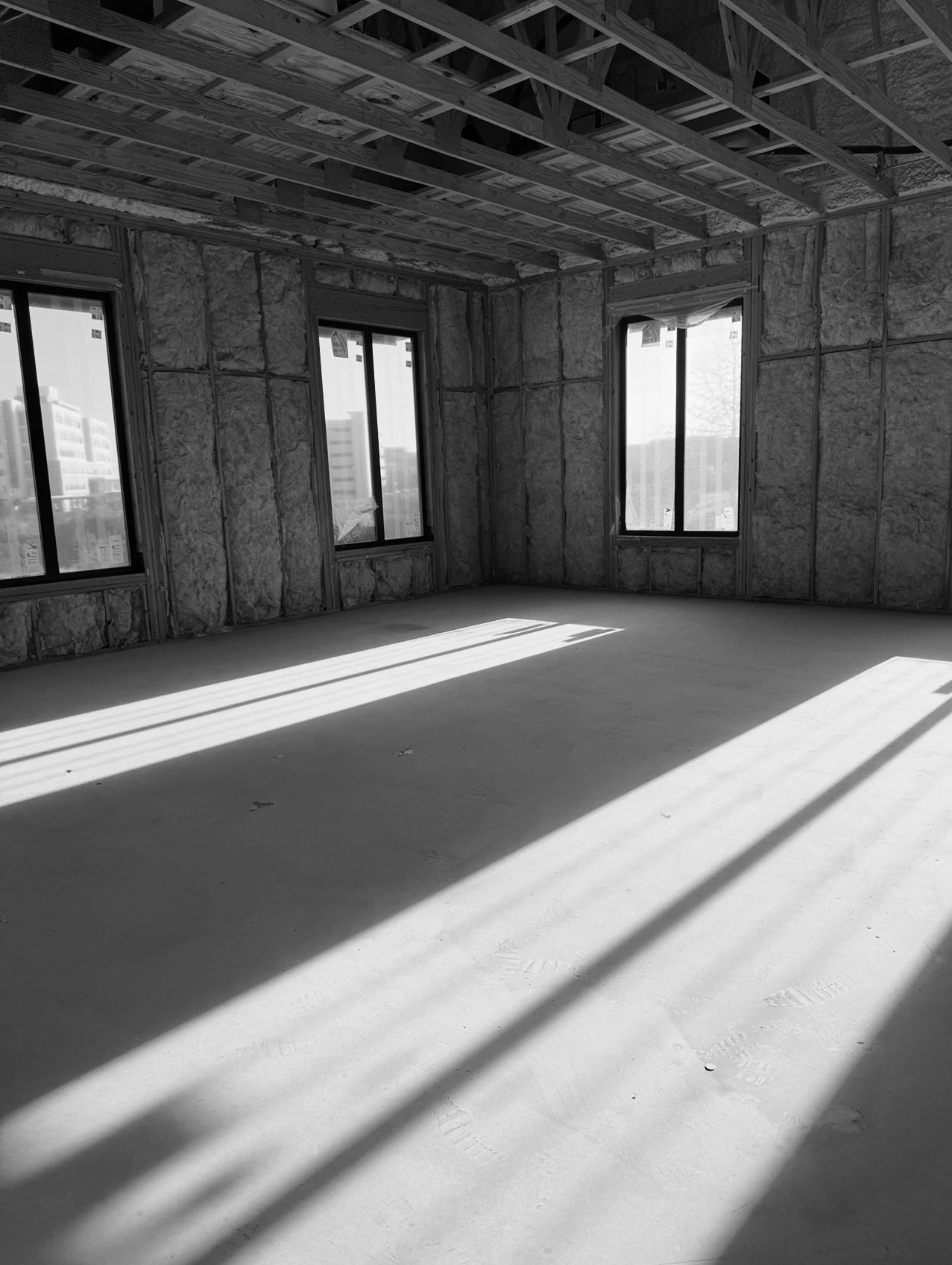Unfinished room in a building under construction with large windows, exposed insulation, and wooden ceiling framing, sunlight casting shadows on the concrete floor.