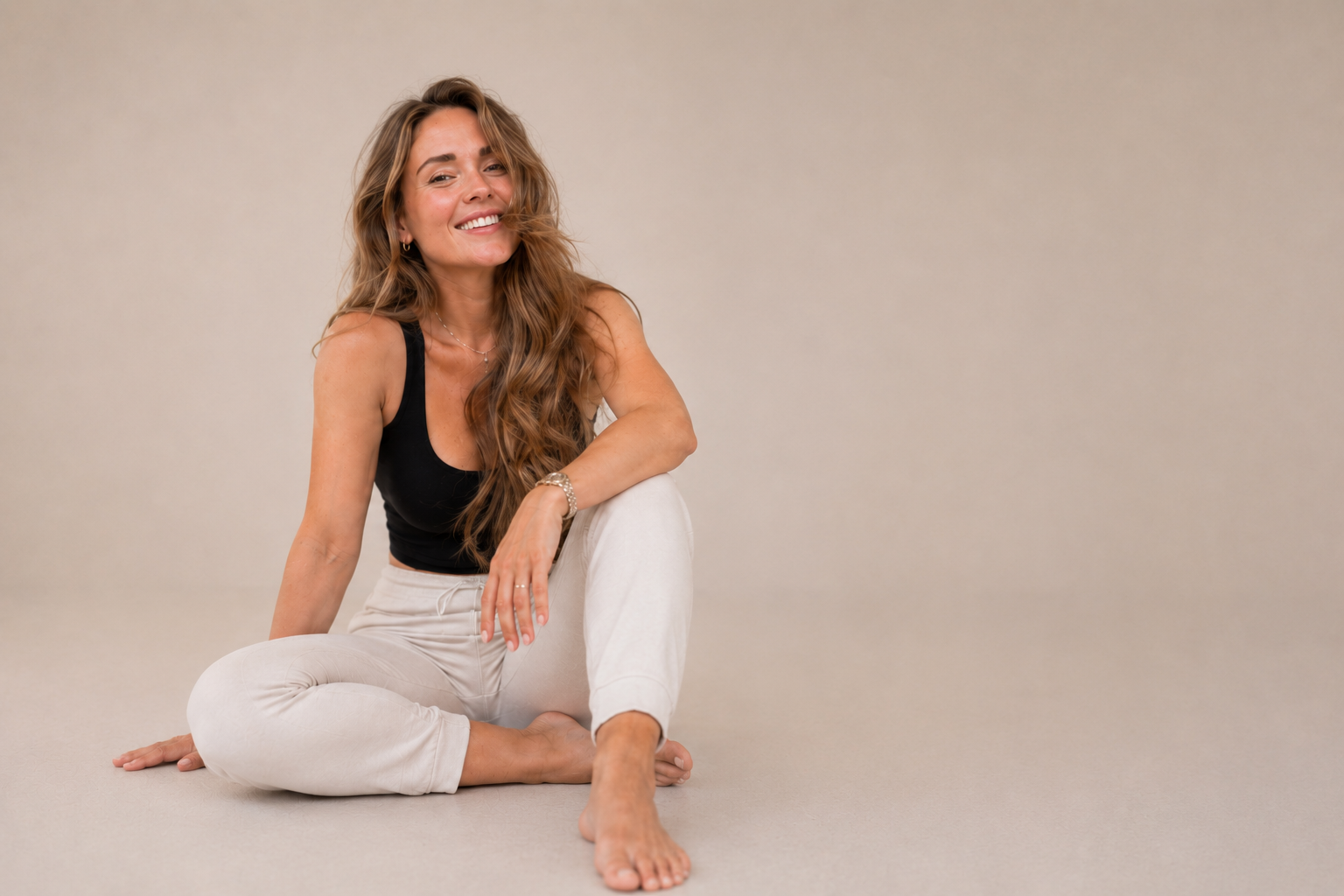 A woman with long wavy hair and a big smile sitting cross-legged on the floor, wearing a black tank top and light-colored pants.