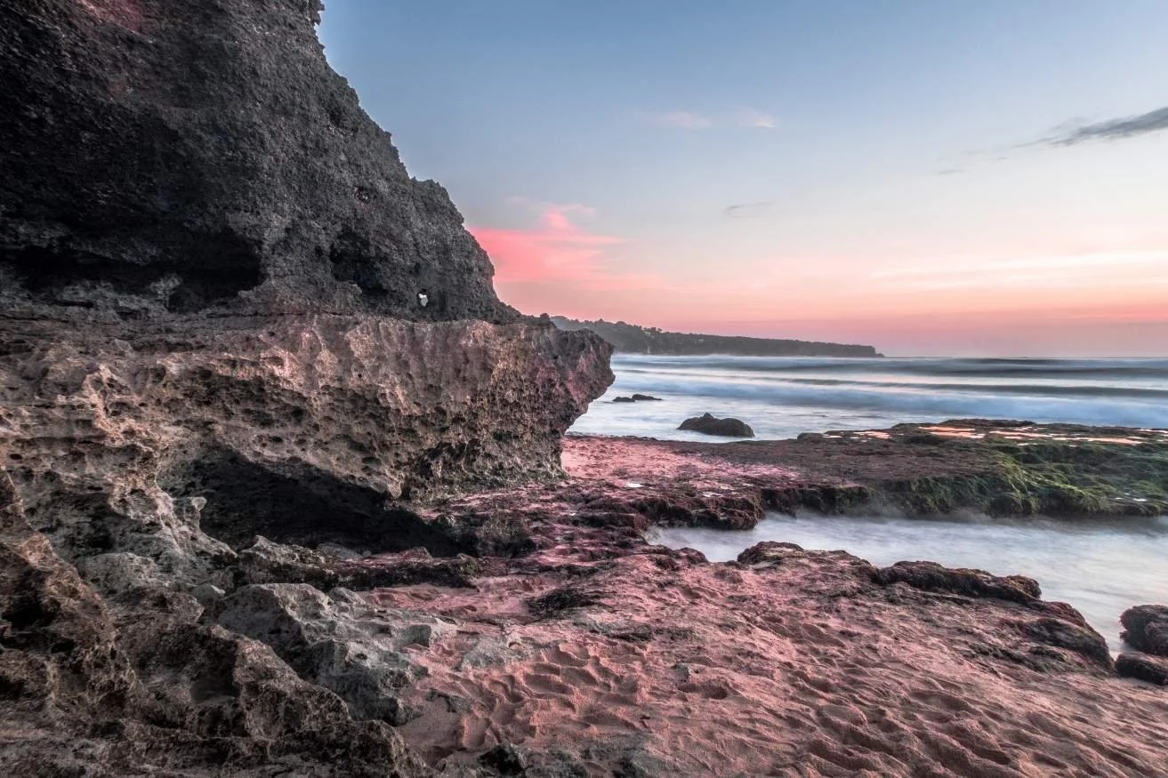 A rocky shoreline at sunset with pink and purple sky, waves crashing against rocks, and a cliff on the left side.