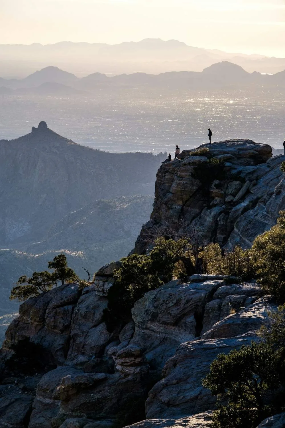 People standing on rocky cliff overlooking a vast landscape with mountains and a shimmering water body in the distance during sunset.