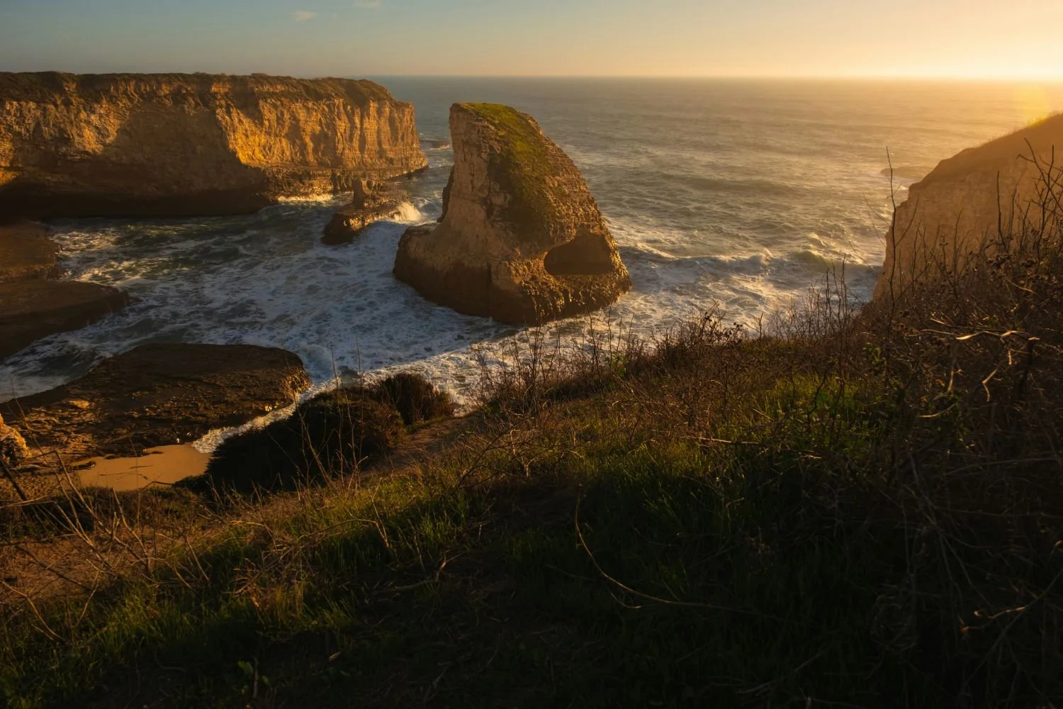 Sunset over a rugged coastline with large rock formations, ocean waves, and sparse vegetation in the foreground.