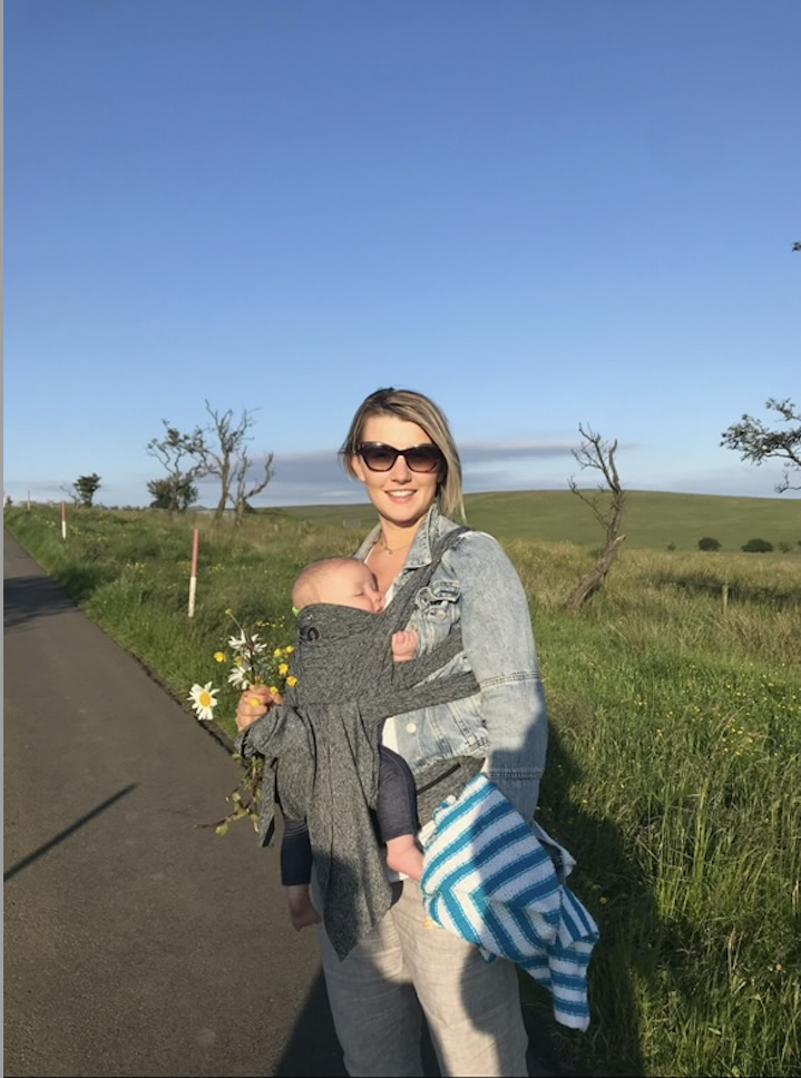 A woman standing outdoors on a sunny day, holding a smiling baby in a carrier on her chest, with a blue sky, grassy fields, and a few scattered trees in the background.