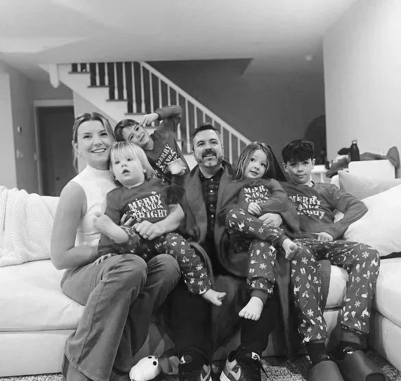 Family of six sitting on a couch in a living room, all wearing matching holiday pajamas, with children and adults smiling.