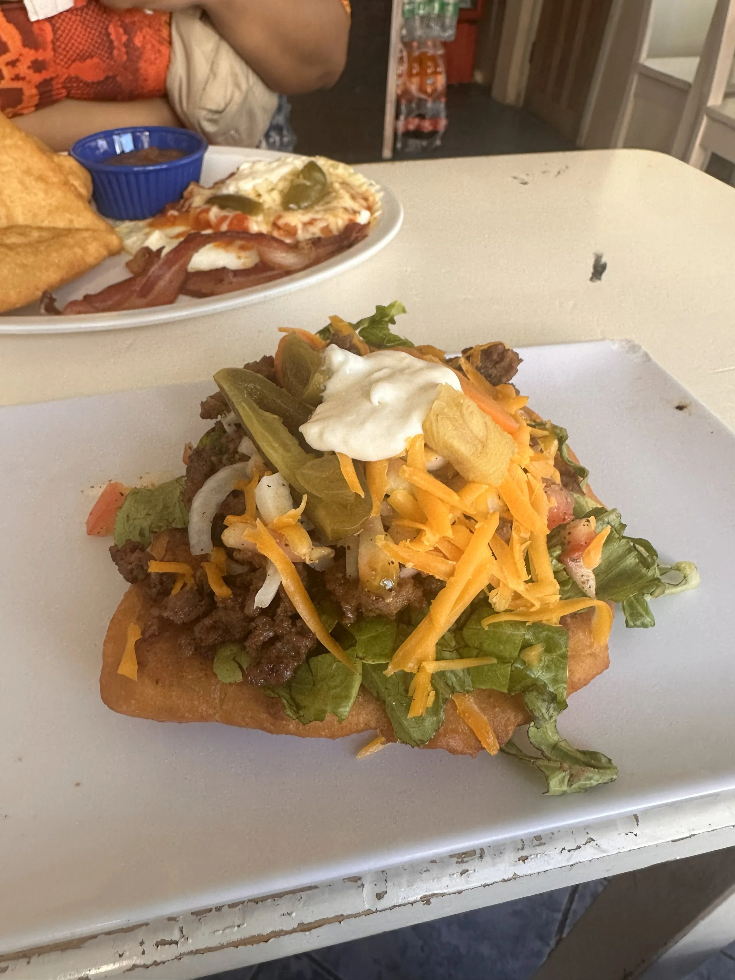 A plate of taco with ground beef, lettuce, shredded cheese, cut jalapeños, sour cream, and a crunchy shell, with a second plate of breakfast food in the background.