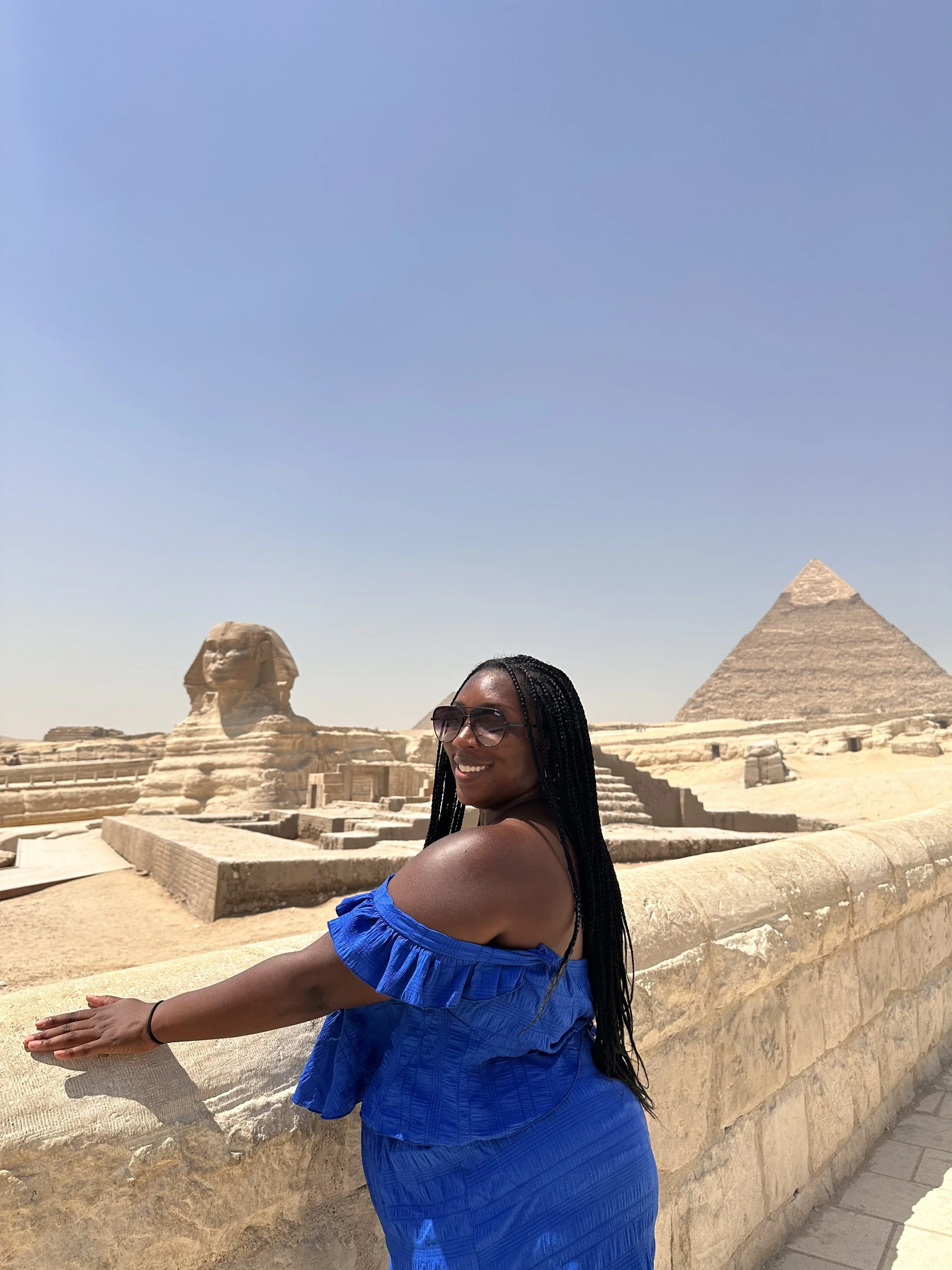 Latrice with long braided hair wearing sunglasses and a blue off-shoulder dress, smiling and leaning on a stone wall at the pyramids and Sphinx in Egypt under a clear blue sky.