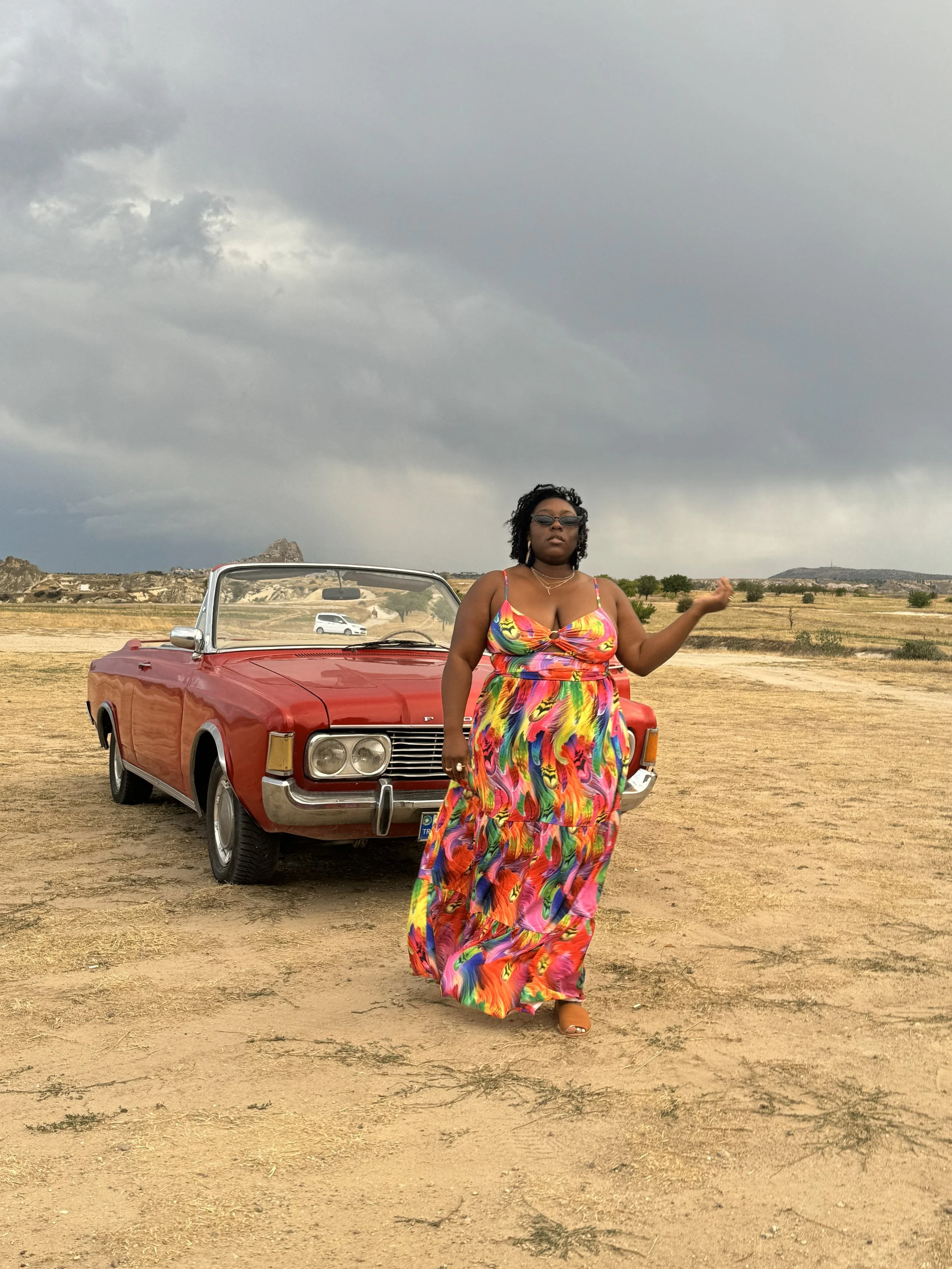 Latrice in a colorful, flowing dress standing in a barren desert with a vintage red convertible car behind her and stormy clouds overhead.