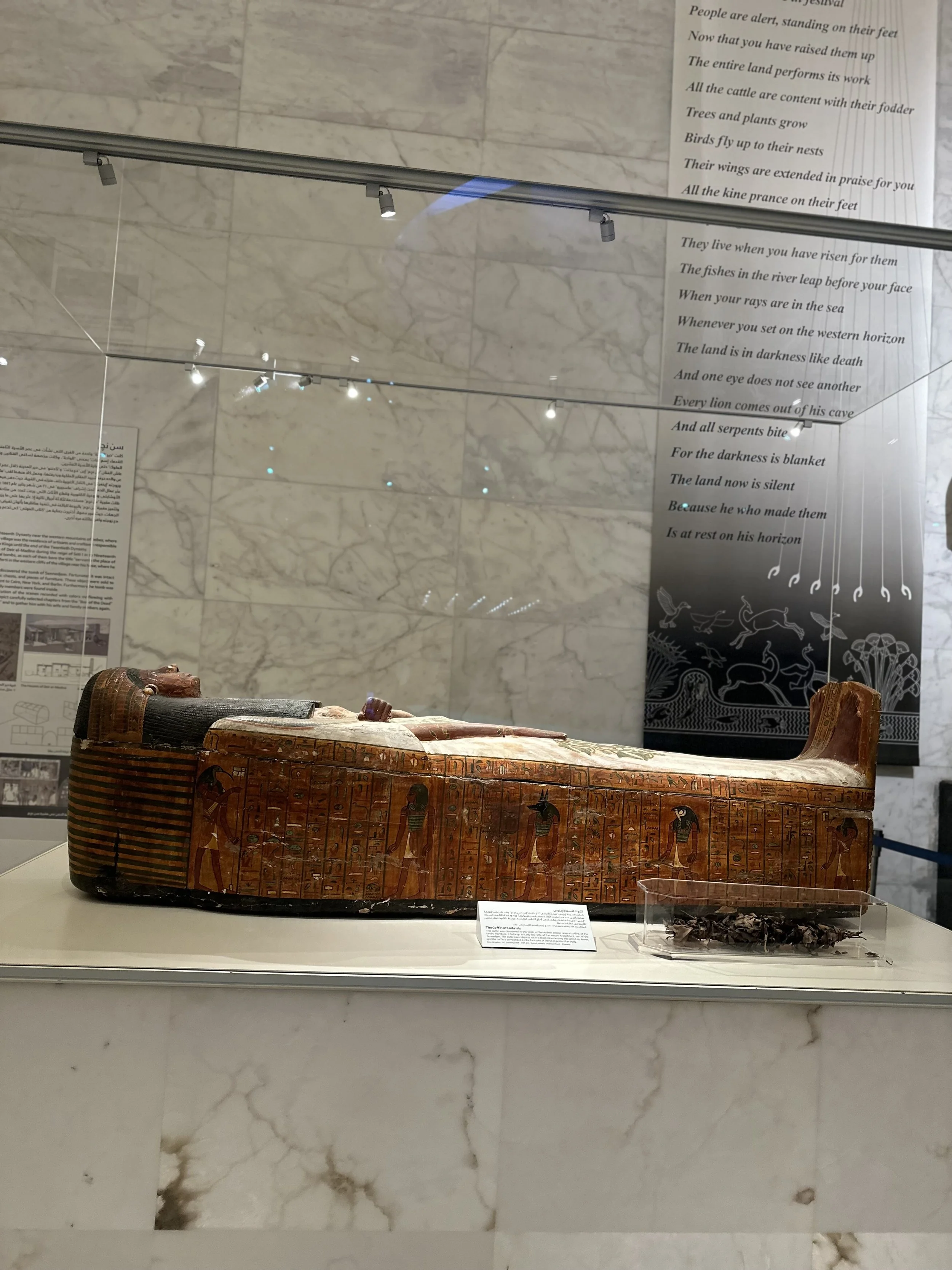 Ancient Egyptian wooden coffin with painted hieroglyphics and figures, displayed on a white marble platform in a museum, with informational signs and a large mural with text in the background.