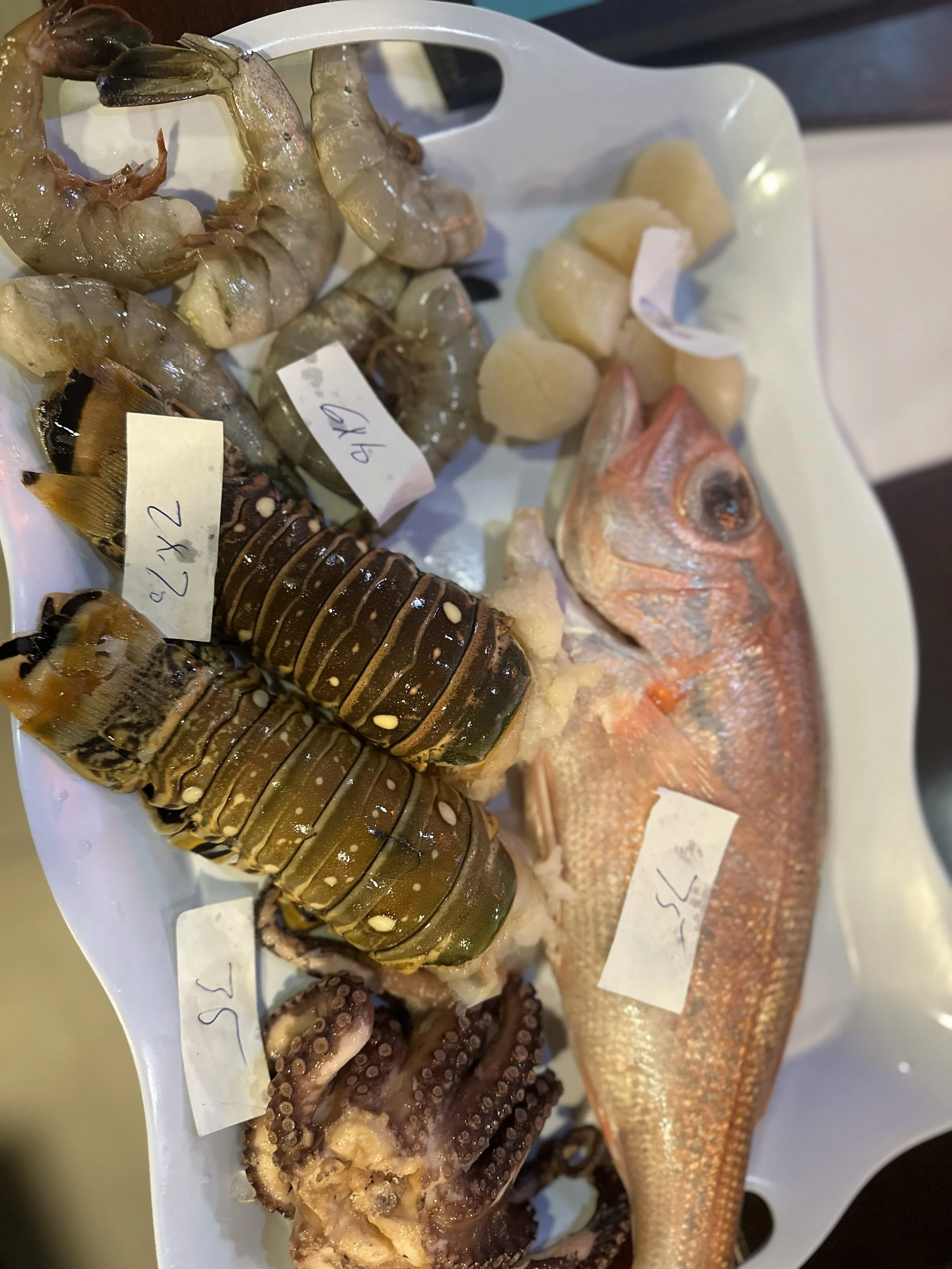 Fresh seafood including shrimp, lobster tail, fish, octopus tentacles, and scallops displayed on a white tray at a market or restaurant.