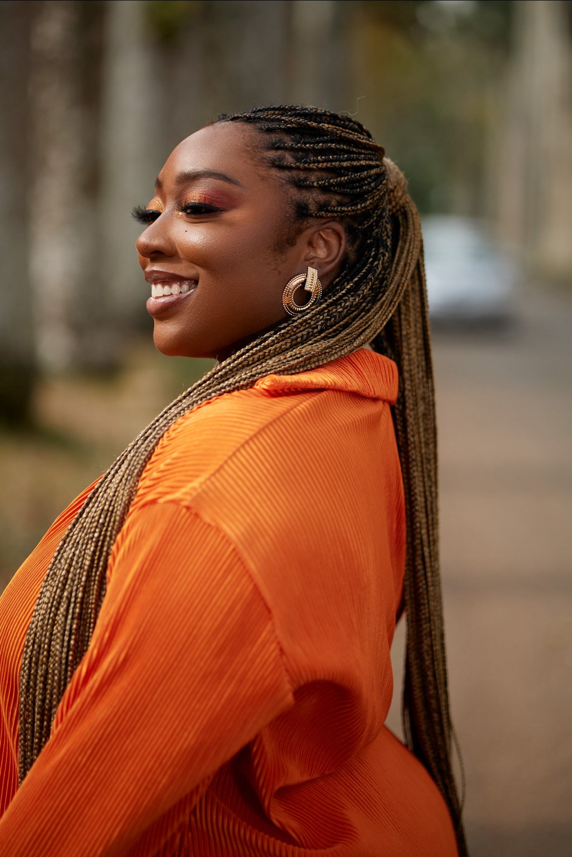 A woman with long, braided hair smiling, wearing a bright orange top and large earrings, outdoors on a blurred street.