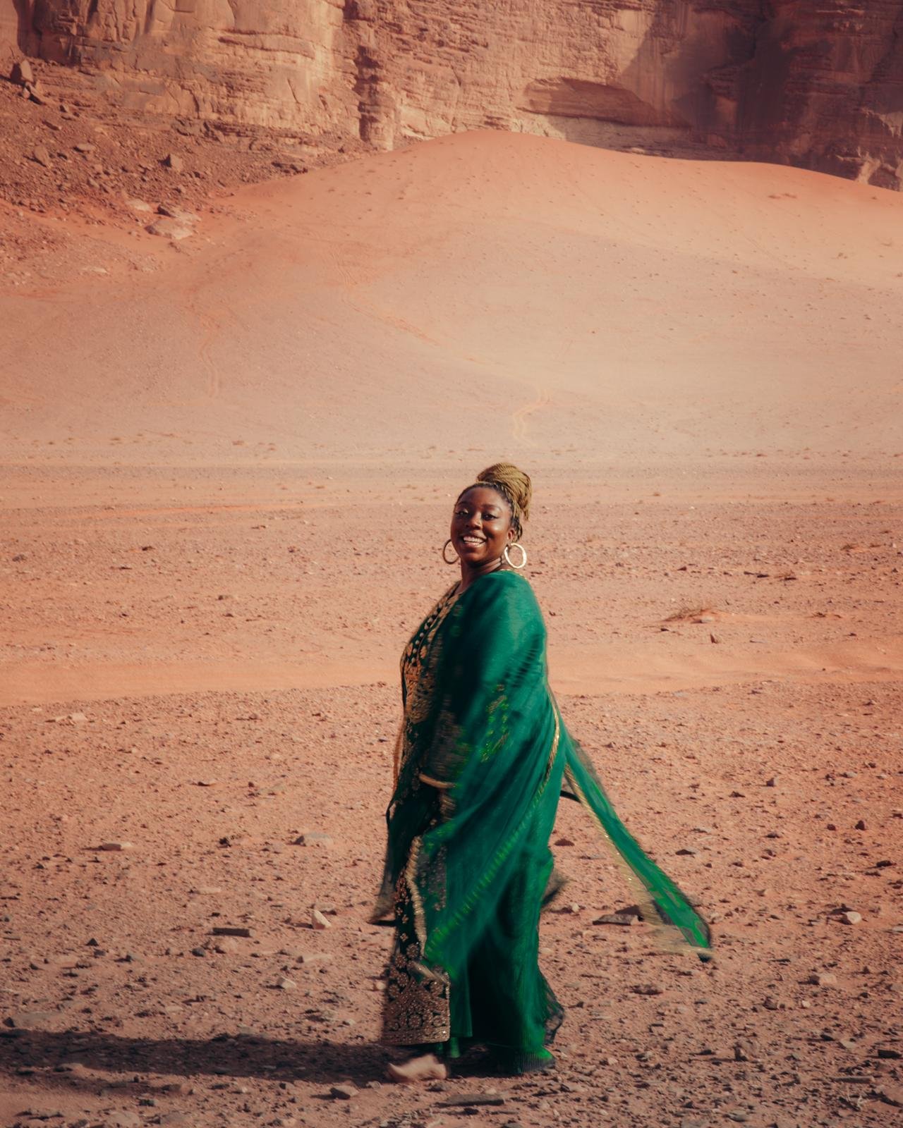 Latrice in a green and black traditional dress standing in a desert landscape with reddish rocks and cliffs in the background, smiling at the camera.