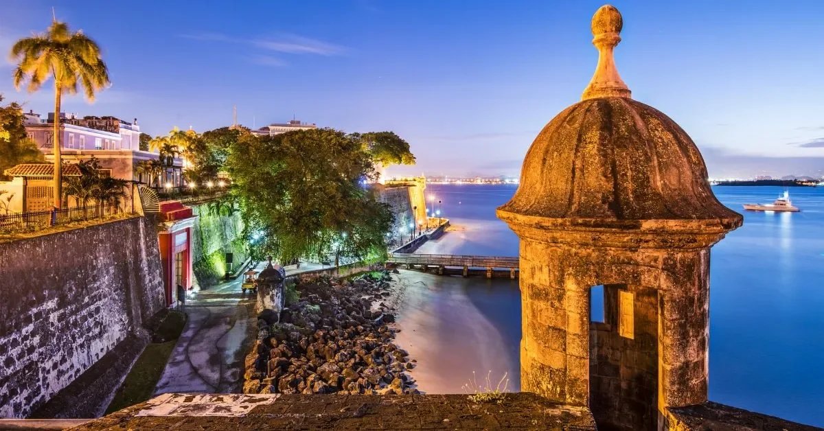 Night view of a historic fortification with a stone sentry tower overlooking a calm body of water, with a large cruise ship in the distance and colorful lights illuminating the shoreline and buildings.