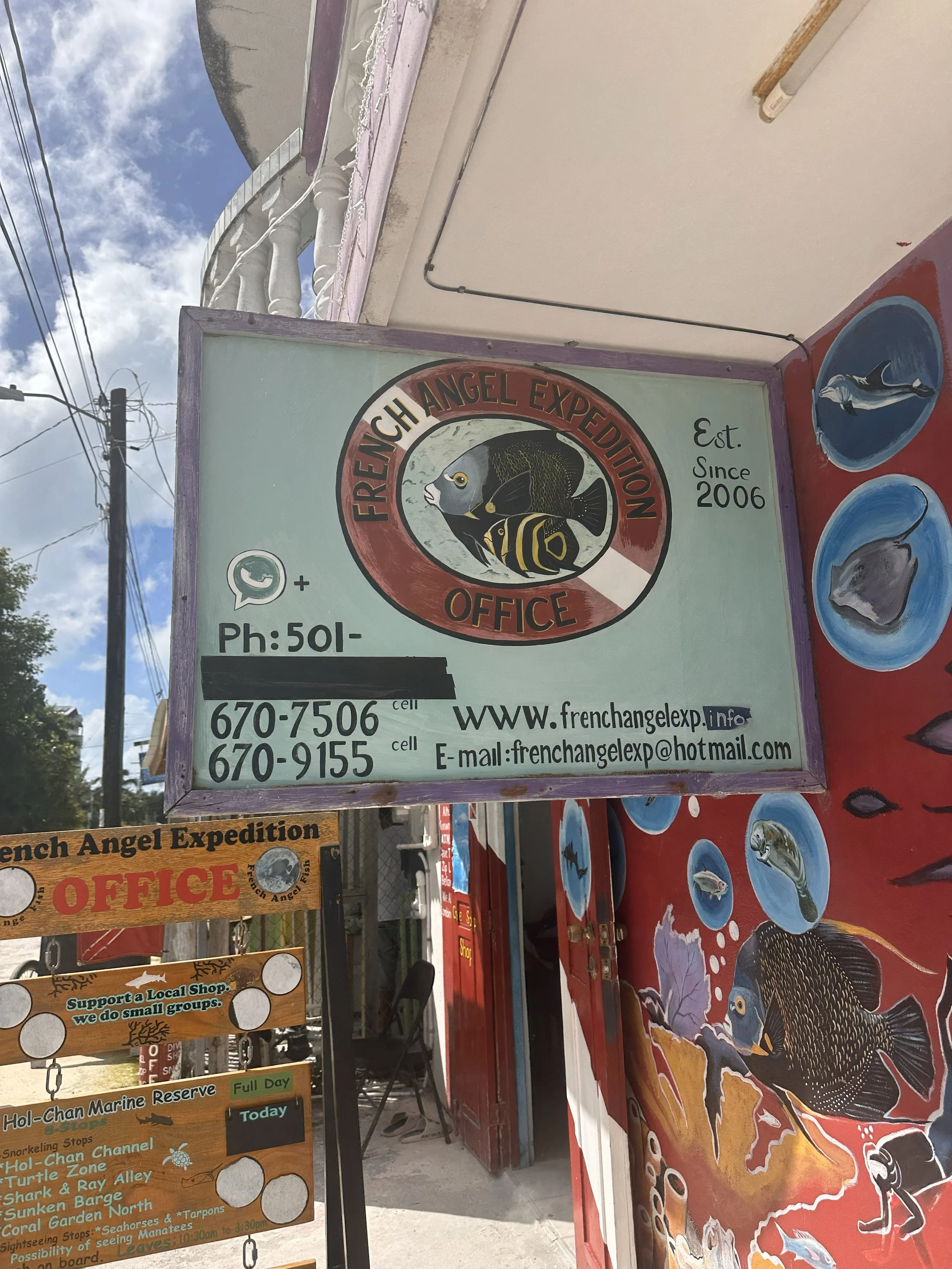 Sign for French Angel Expedition office, established in 2006, with a fish logo, contact phone numbers, website, and email address. The sign is surrounded by regional fishing symbols and colorful illustrations of fish on a red background.