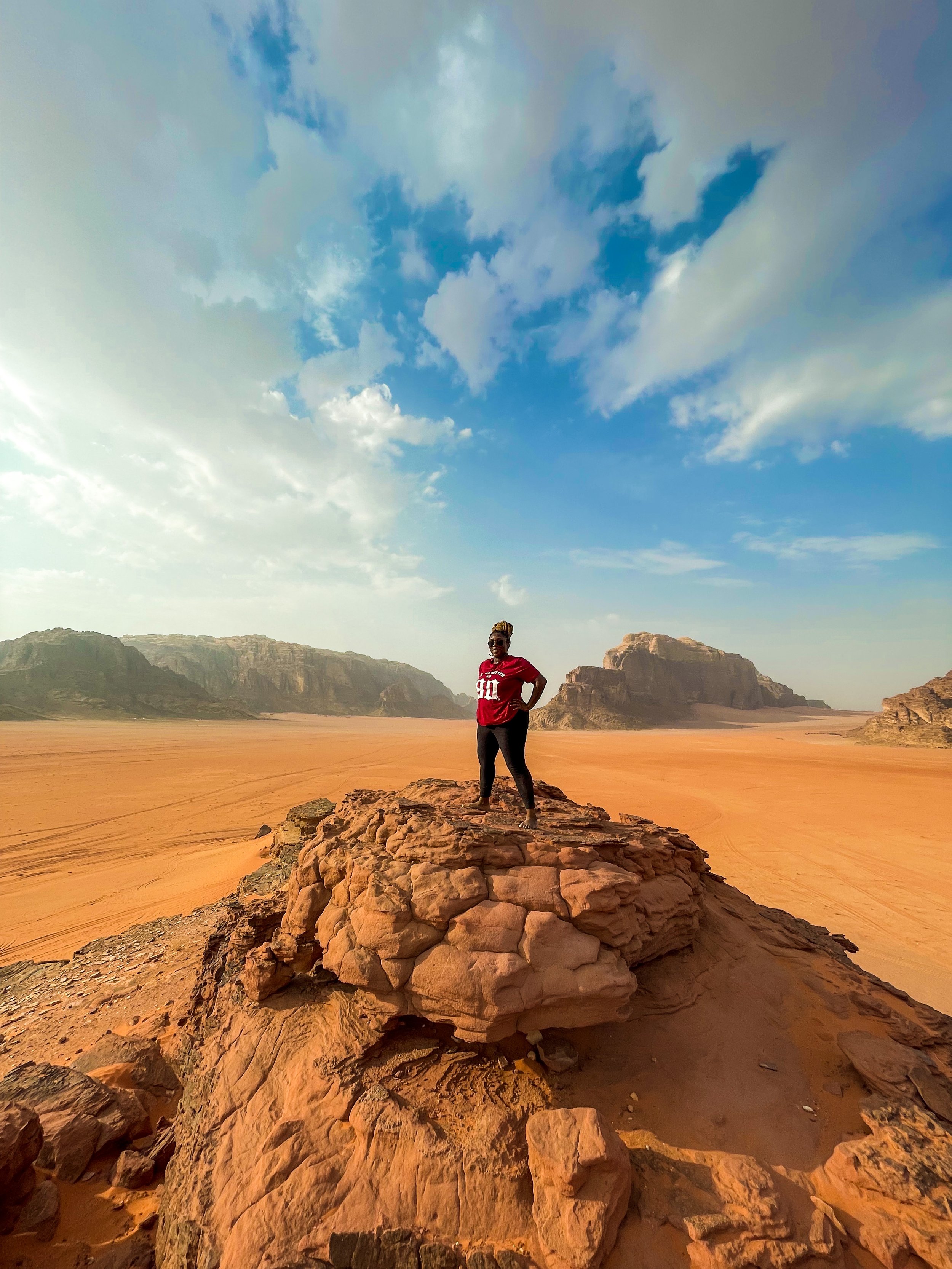 Latrice standing on a large rock formation in a desert landscape with rocky mountains in the background and a partly cloudy sky overhead.