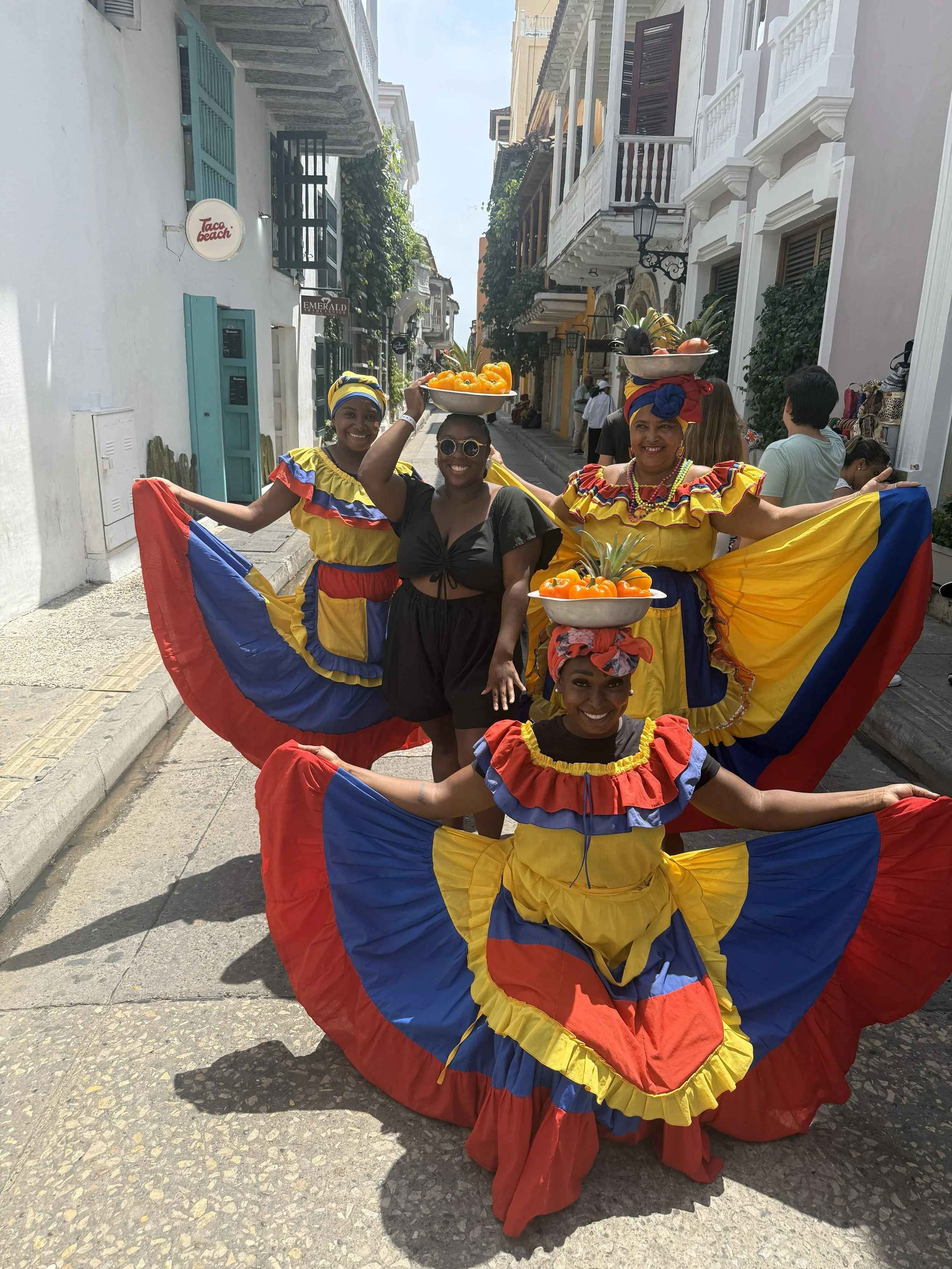Four women in colorful traditional dresses celebrate in a street with colonial-style buildings; three women wear yellow, blue, and red dresses with matching headscarves, holding bowls of fruit on their heads, while one woman in black stands among the