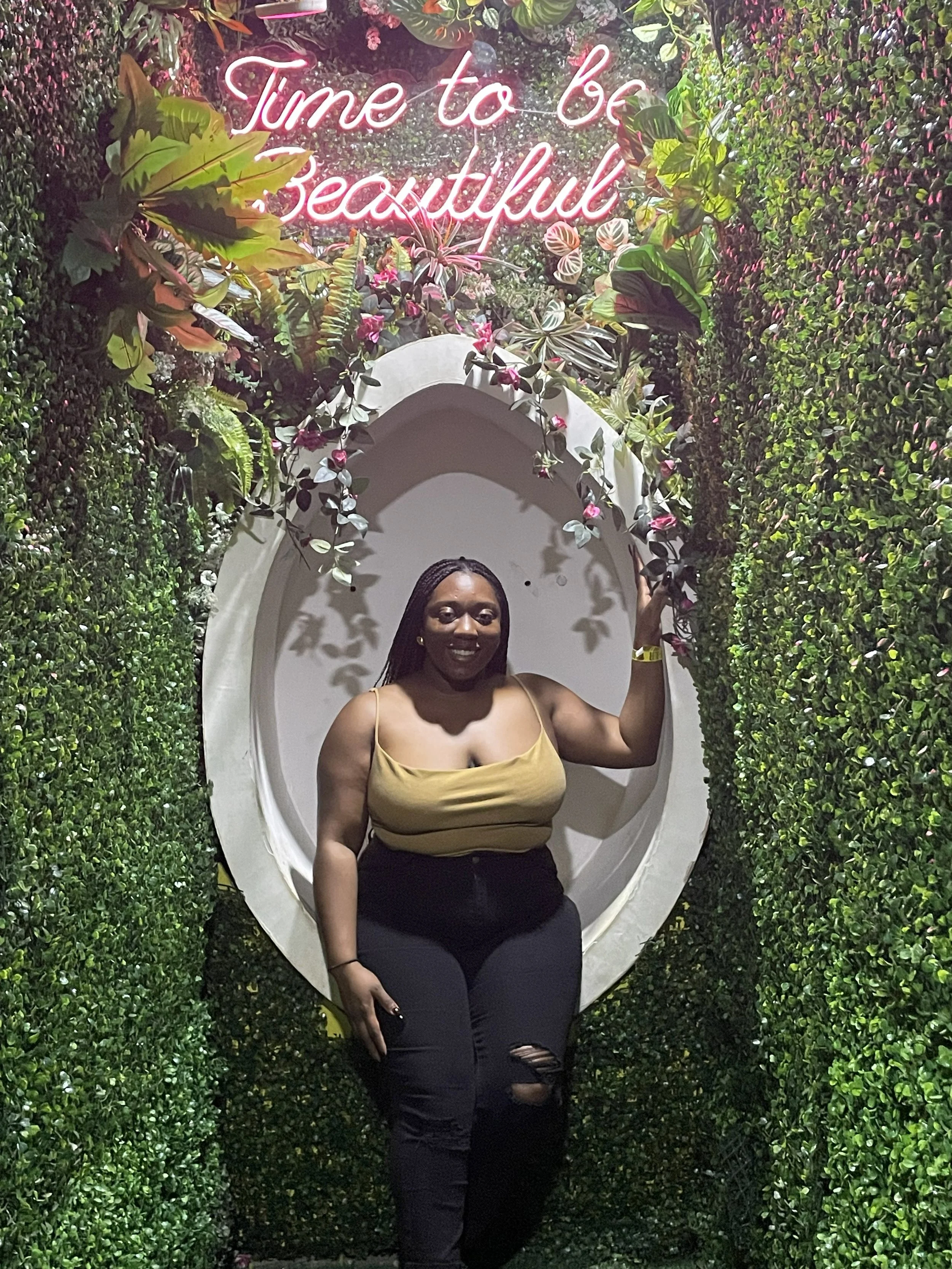 Latrice in a yellow top and ripped black jeans posing in front of a decorative wall with a neon sign that says 'Time to be Beautiful' and a floral and greenery backdrop.