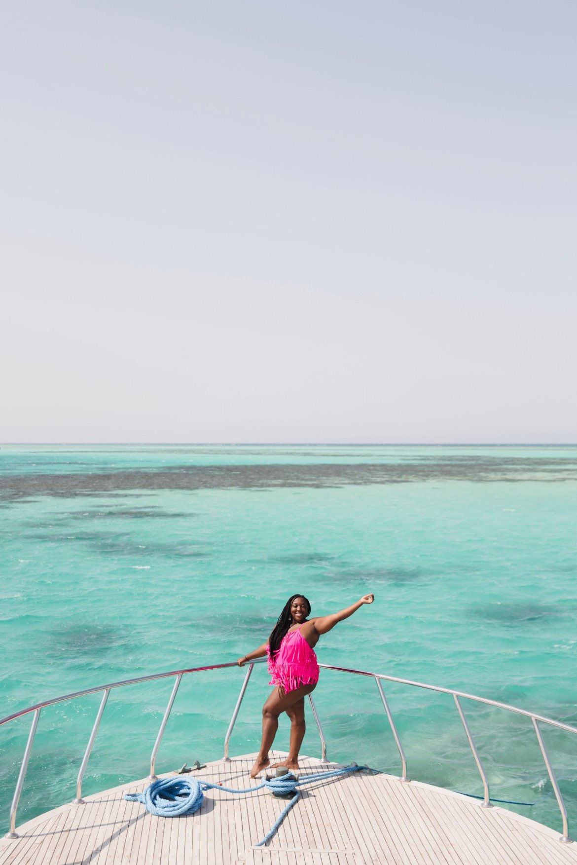 A woman in a pink dress standing barefoot on the deck of a boat, posing with her arm extended outward, surrounded by turquoise ocean water under a cloudy sky.