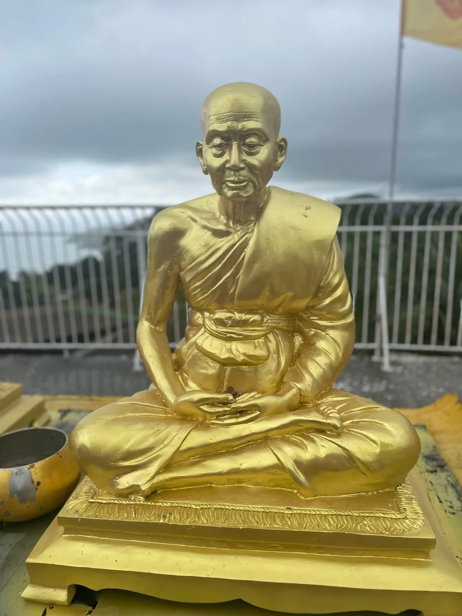 A gold-colored statue of an elderly man in traditional clothing, sitting cross-legged with hands resting in his lap, on a decorative base. The background shows a cloudy sky and a metal fence.