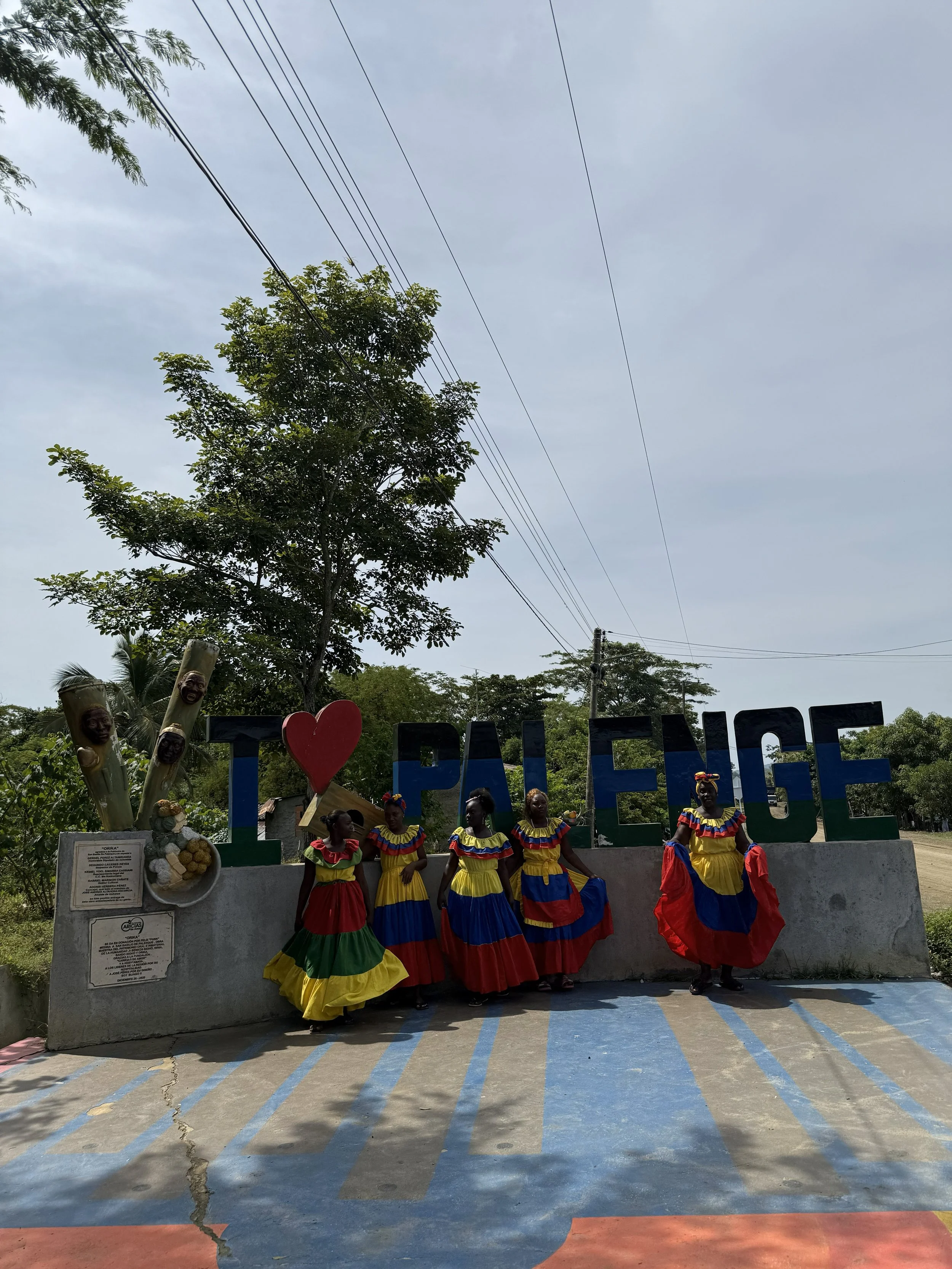 A large sign reading "I Love Paeng" with four women in colorful traditional dresses standing in front of it. There are trees and power lines in the background.