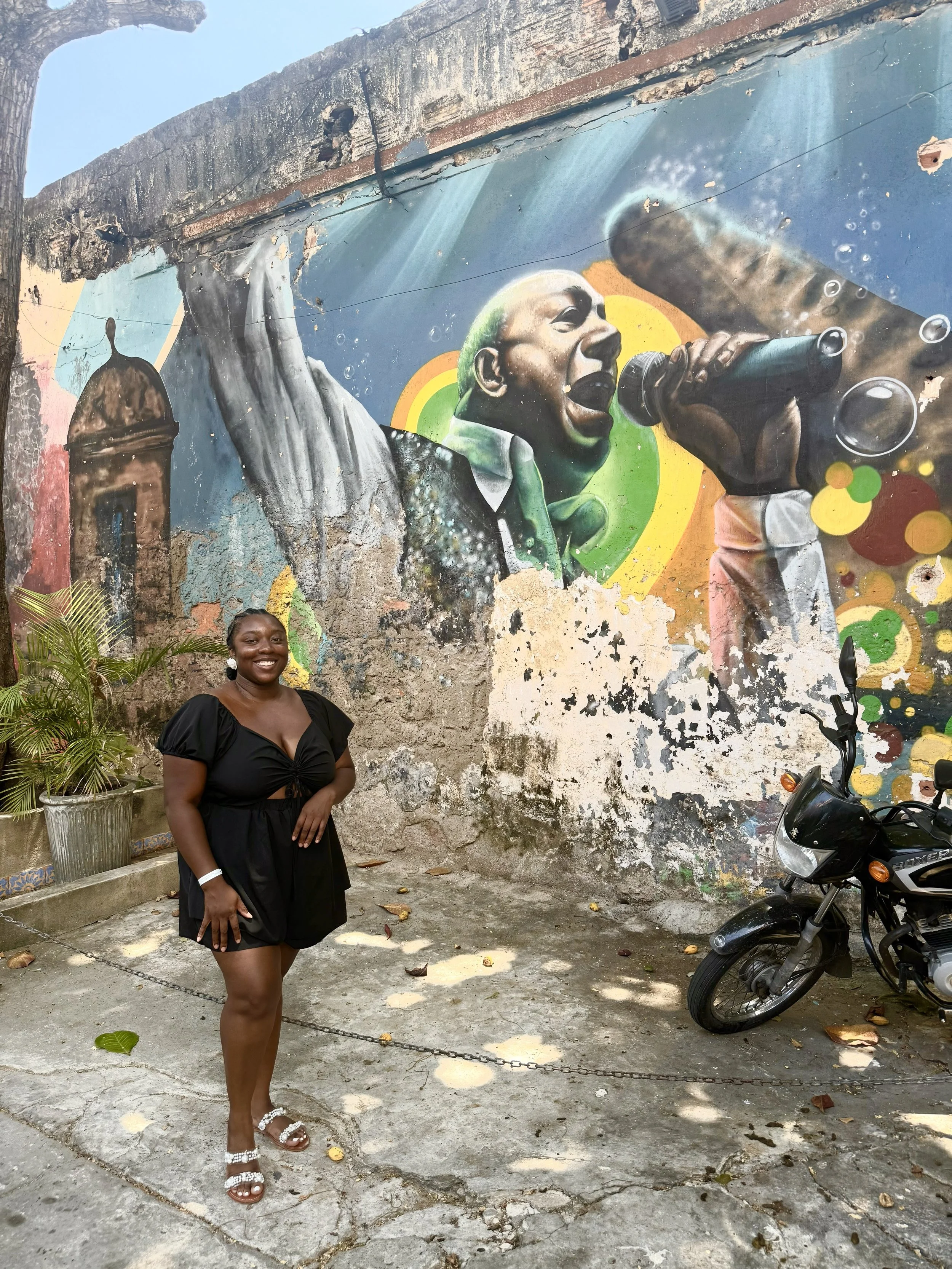 Latrice in a black dress smiling and standing in front of a colorful street art mural of a man singing into a microphone. There is a motorcycle to the right and potted plants on the left.